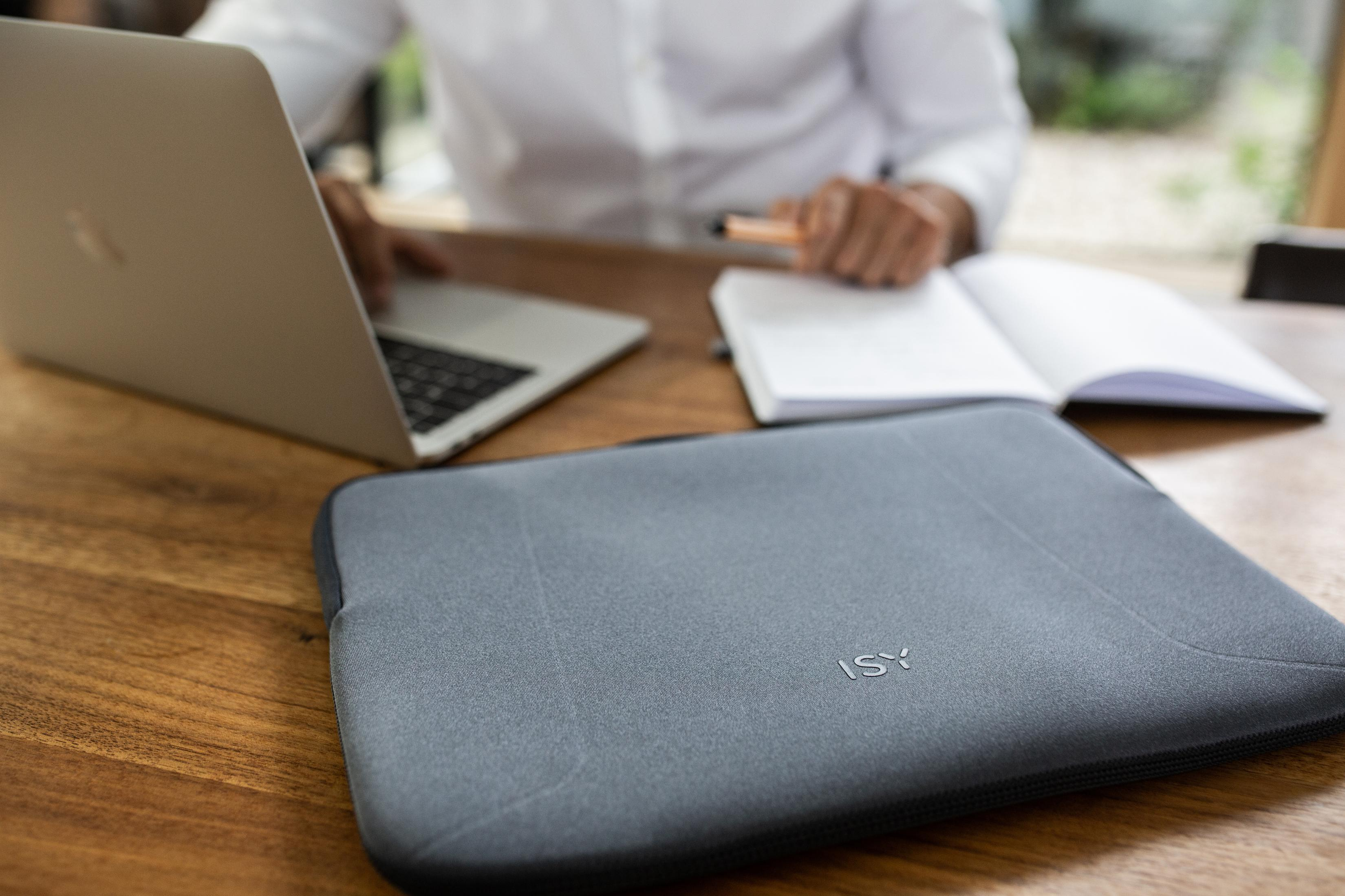 Laptop auf einem Schreibtisch. Hände einer Person sind in der Nähe des Laptops. Ein offenes Notizbuch und ein Stift liegen ebenfalls auf dem Schreibtisch.