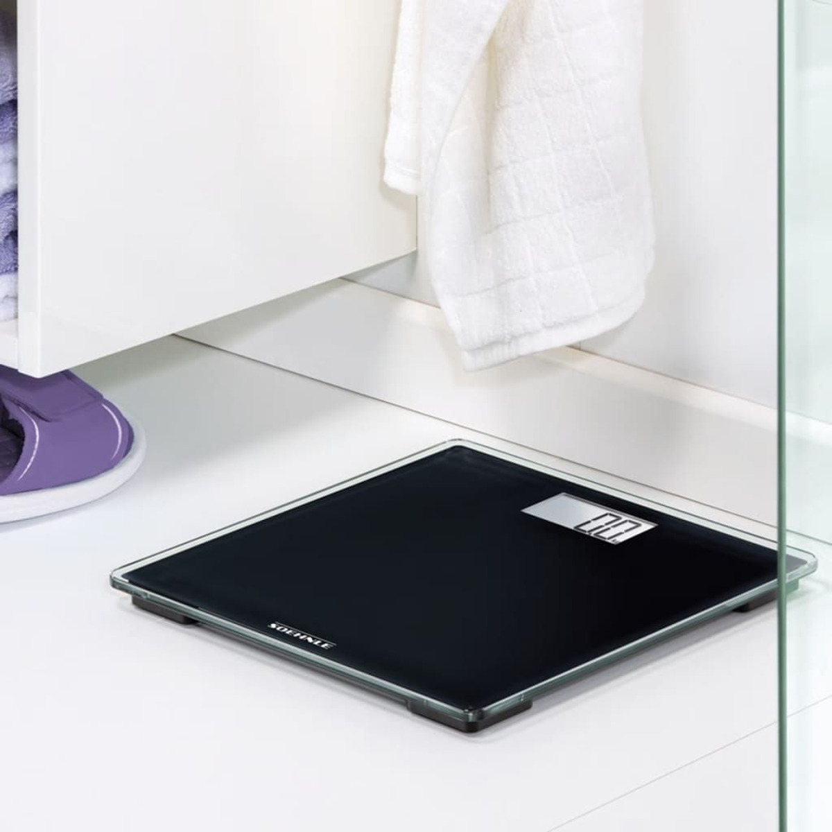 Schwarze Digitalwaage auf weißem Fliesenboden, mit weißem Handtuch und Schrank im Hintergrund.