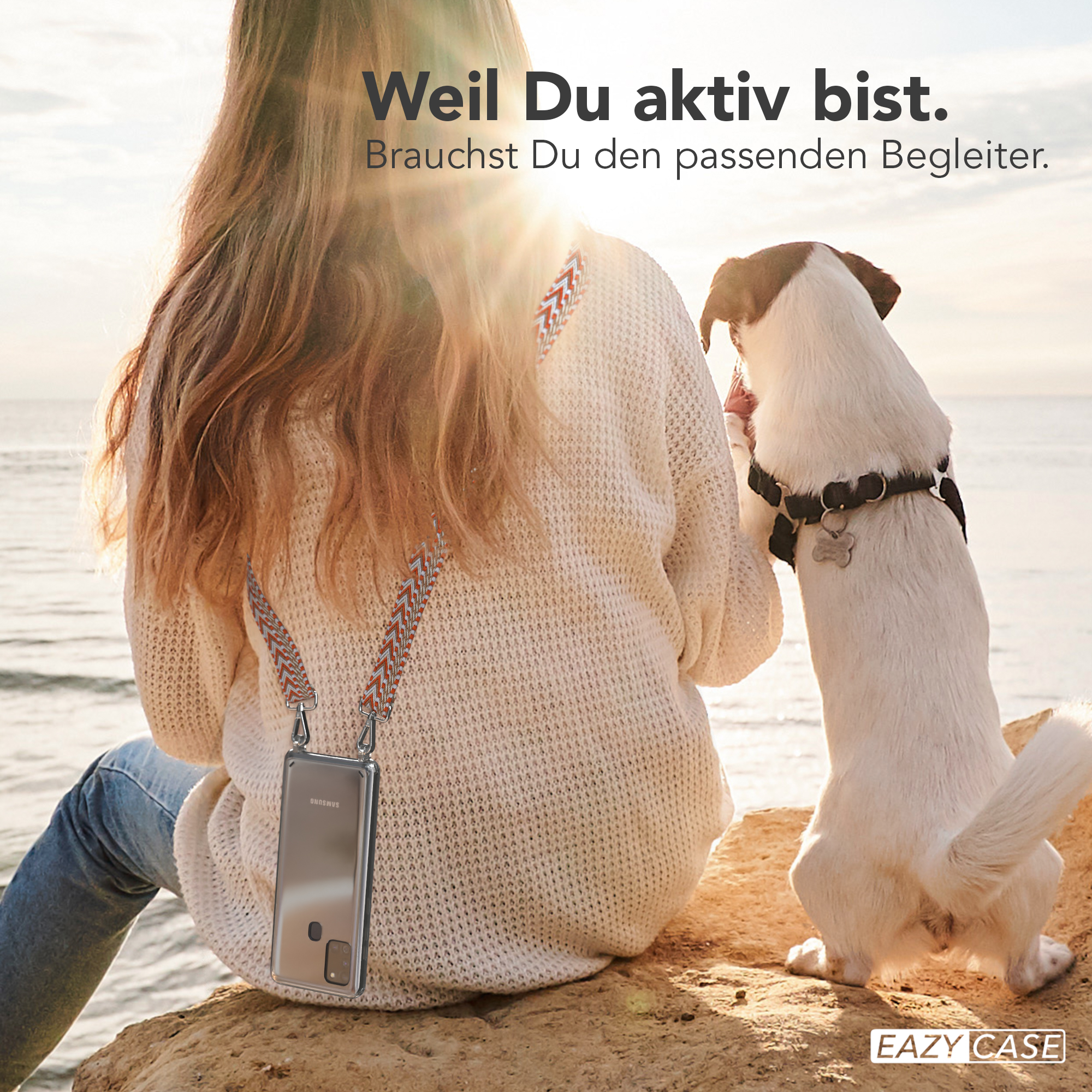 Frau mit Hund am Strand. Sie schauen auf ein Handy in einer orange-grauen Hülle. Die Sonne scheint.