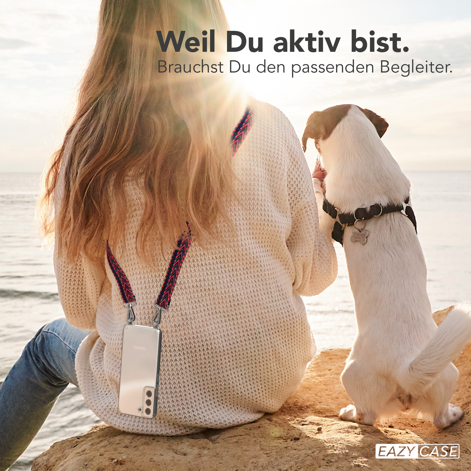 Frau mit Telefon am Riemen, Hund beobachtet. Sonne scheint im Hintergrund.