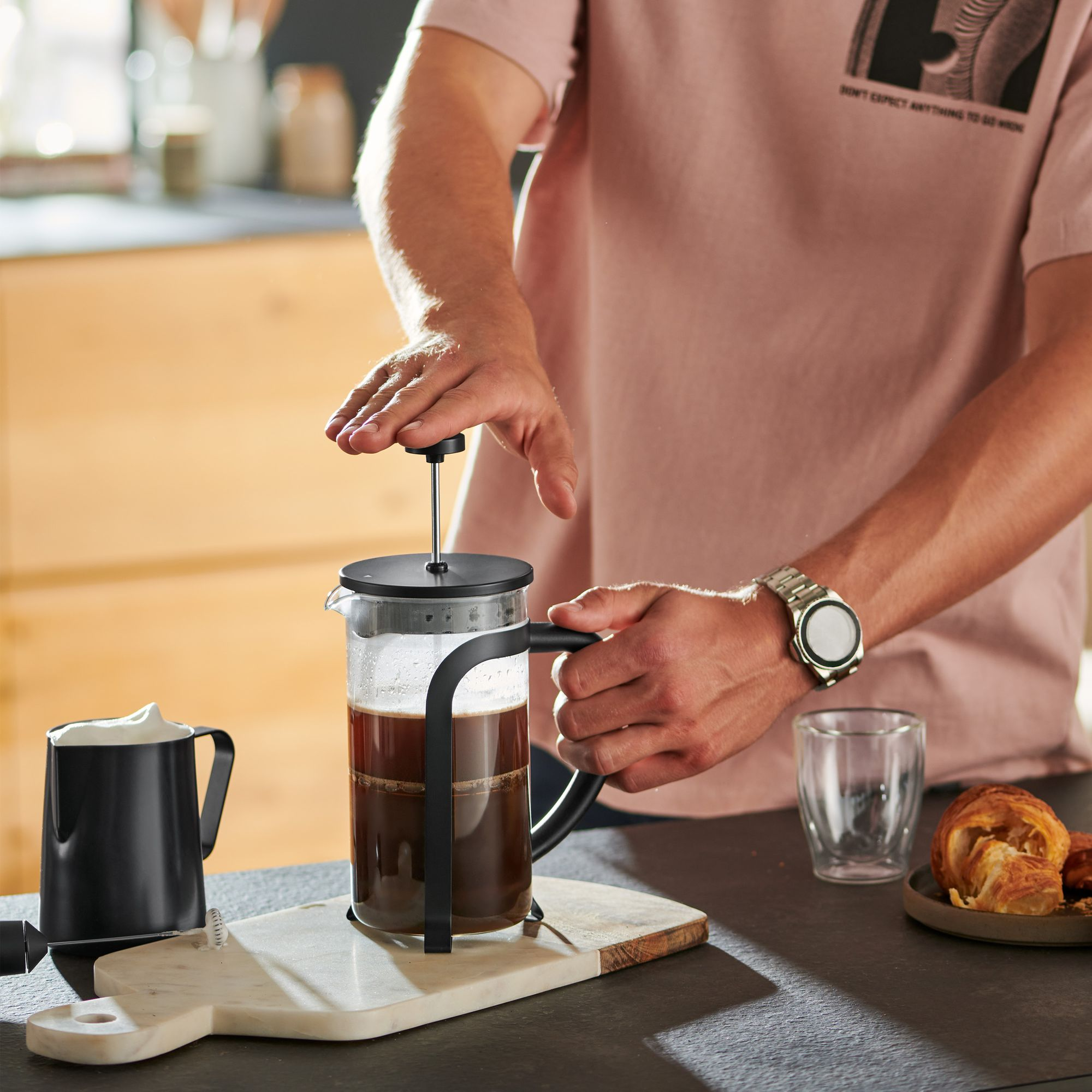 Person benutzt eine French Press. Kaffee im Glas, eine schwarze Tasse und ein Croissant auf der Theke.