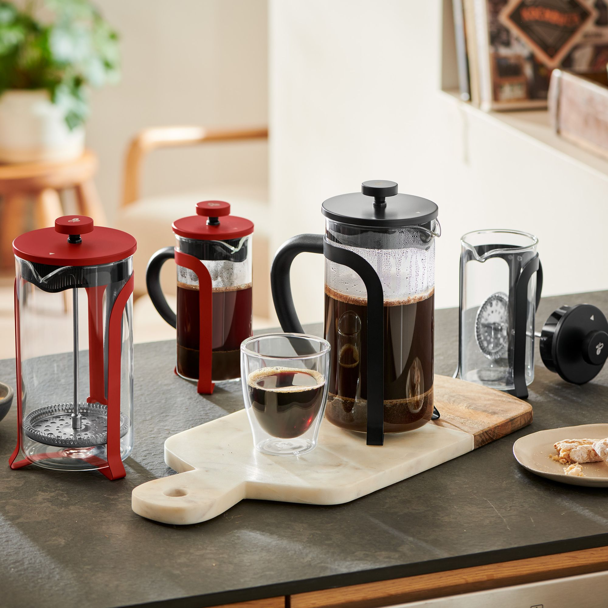 Drei French Presses und ein Glas Kaffee auf einer Theke. Der Kaffee ist dunkel.
