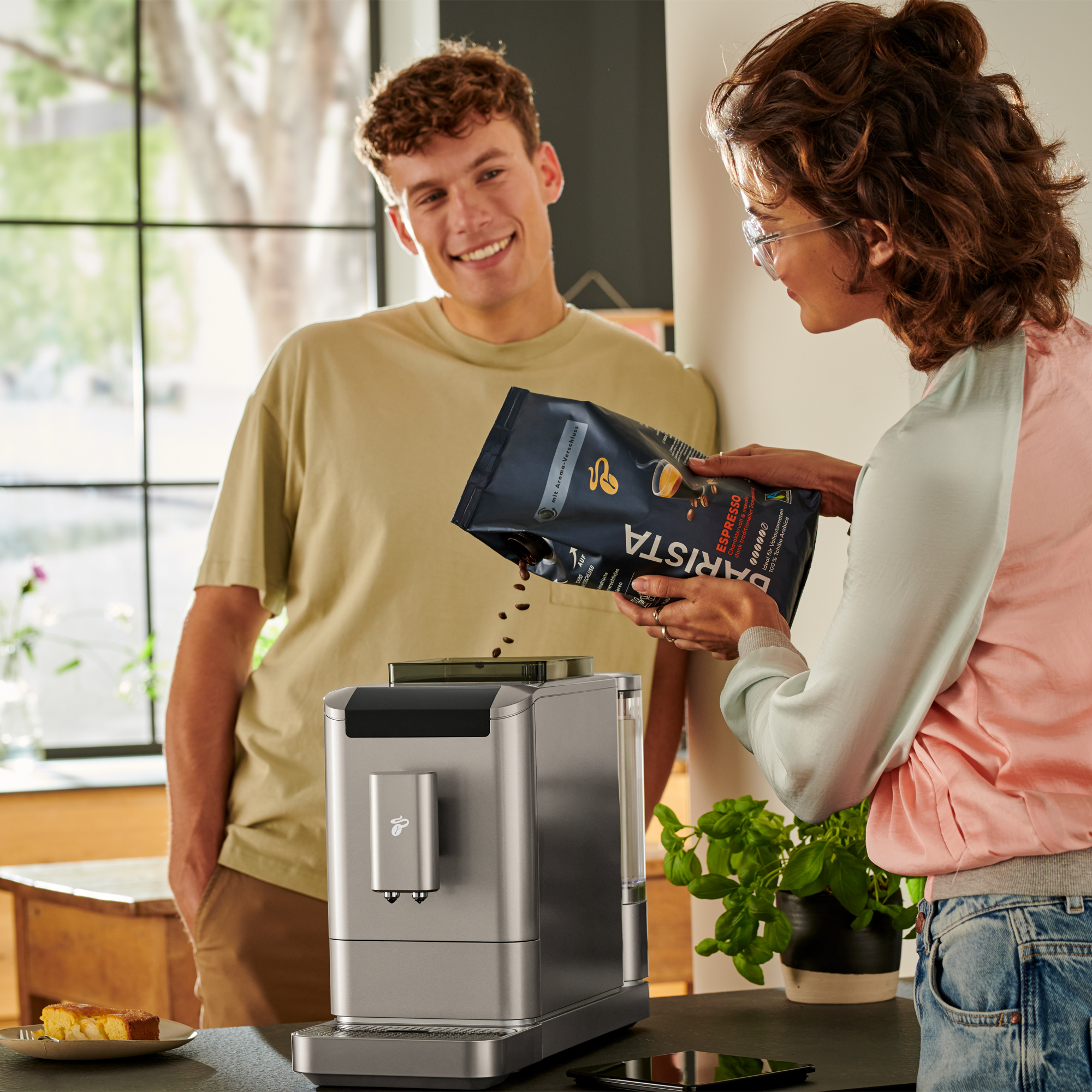 Paar mit Kaffeemaschine, einer gießt Bohnen in die Maschine.