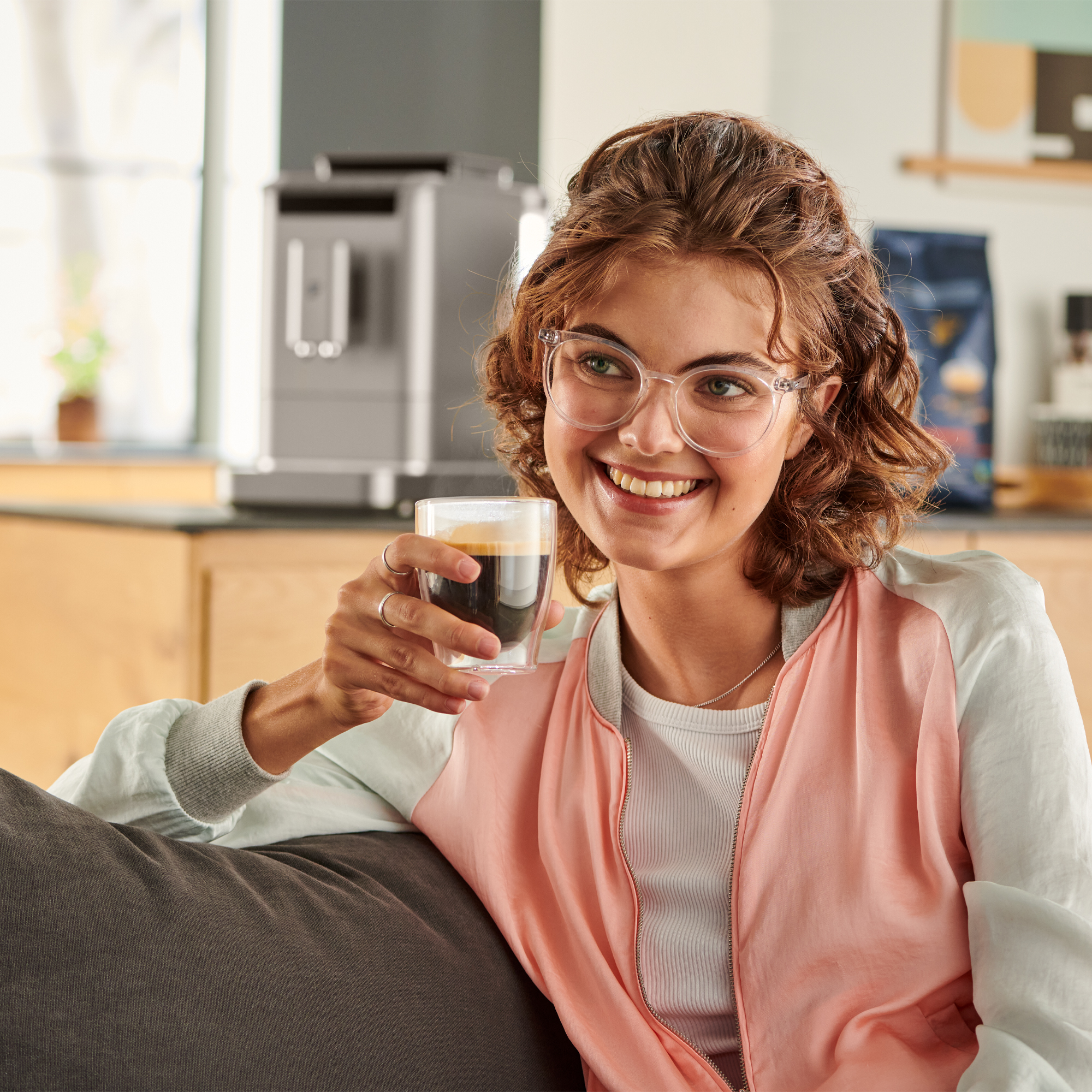 Eine Frau mit Brille lächelt, während sie ein Glas Kaffee hält, mit einer Kaffeemaschine und einer Tüte Kaffeebohnen.