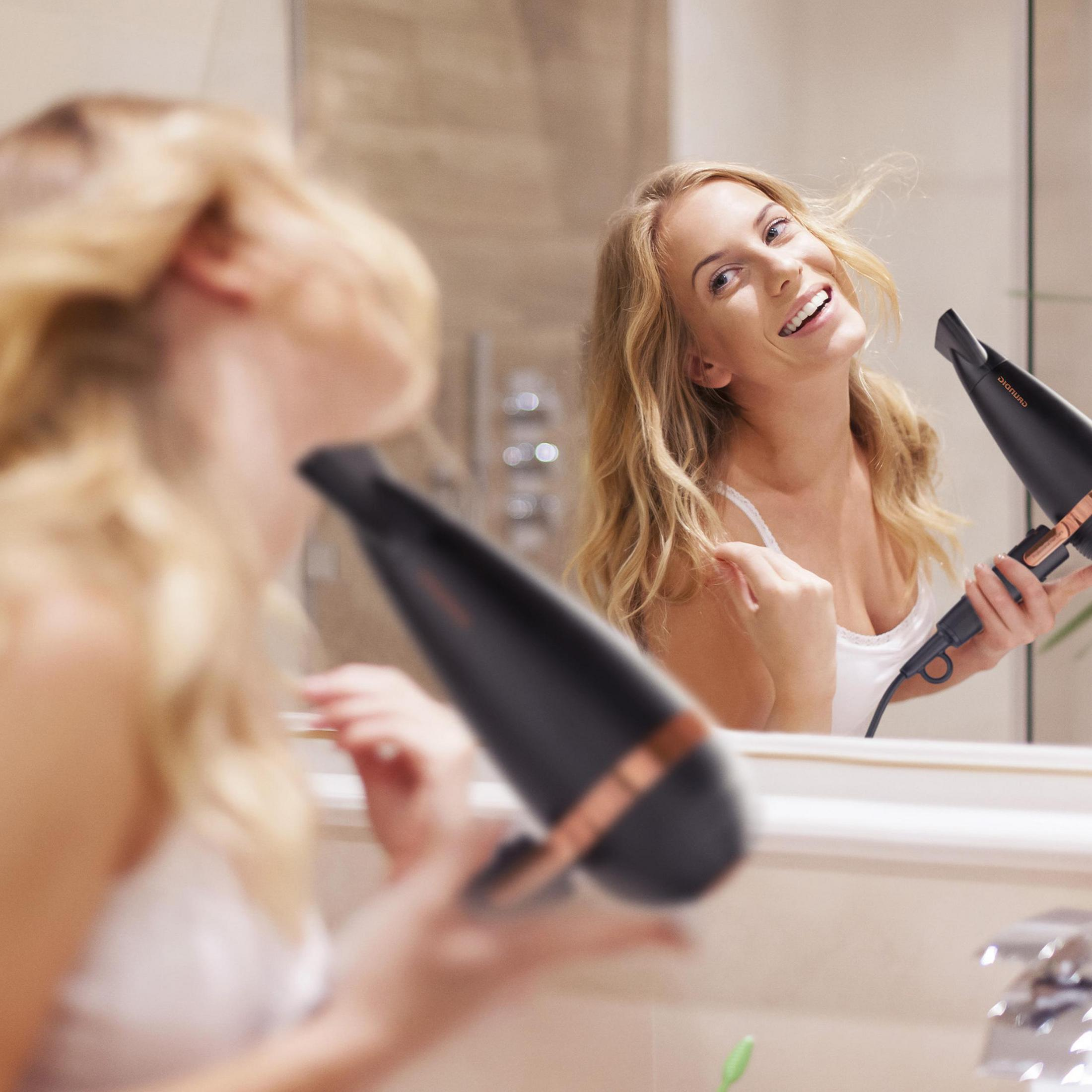 Kobieta susząca włosy przed lustrem, uśmiechnięta z czarną suszarką w dłoni.