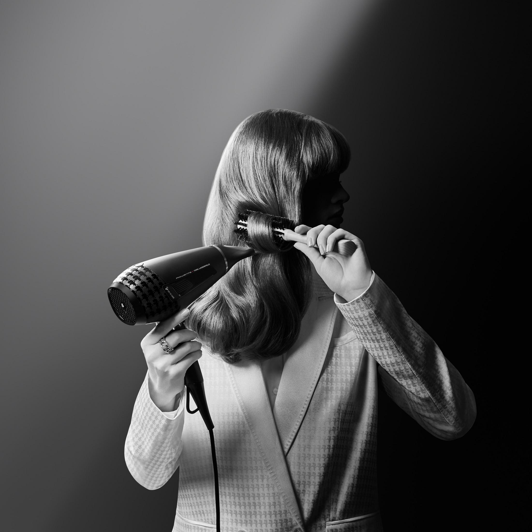 Kobieta stylizująca włosy suszarką i szczotką, zdjęcie czarno-białe.