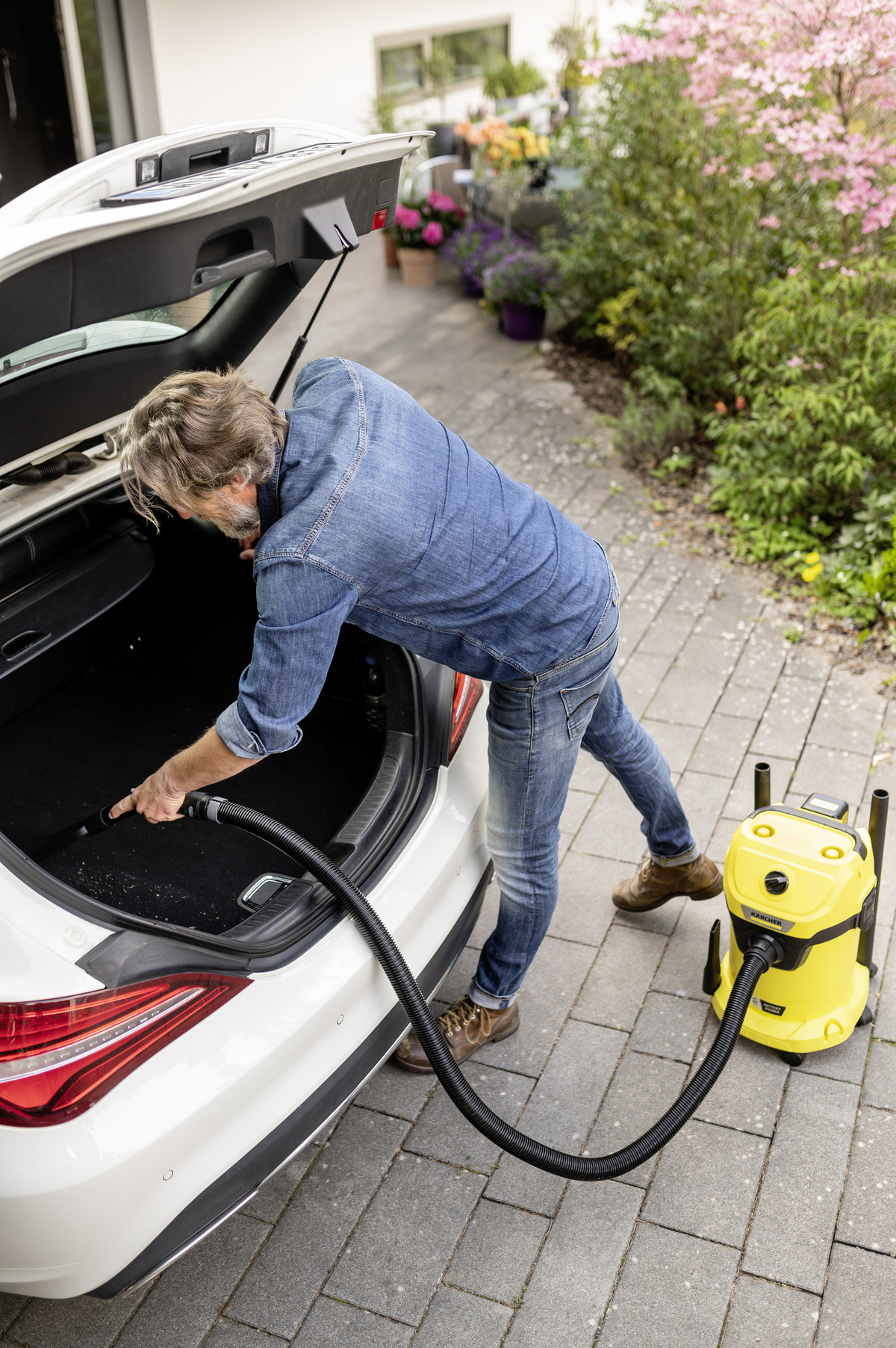 Ein Mann saugt den Kofferraum eines weißen Autos. Ein gelber Staubsauger steht auf dem Boden. Er trägt Jeans und ein Jeanshemd.