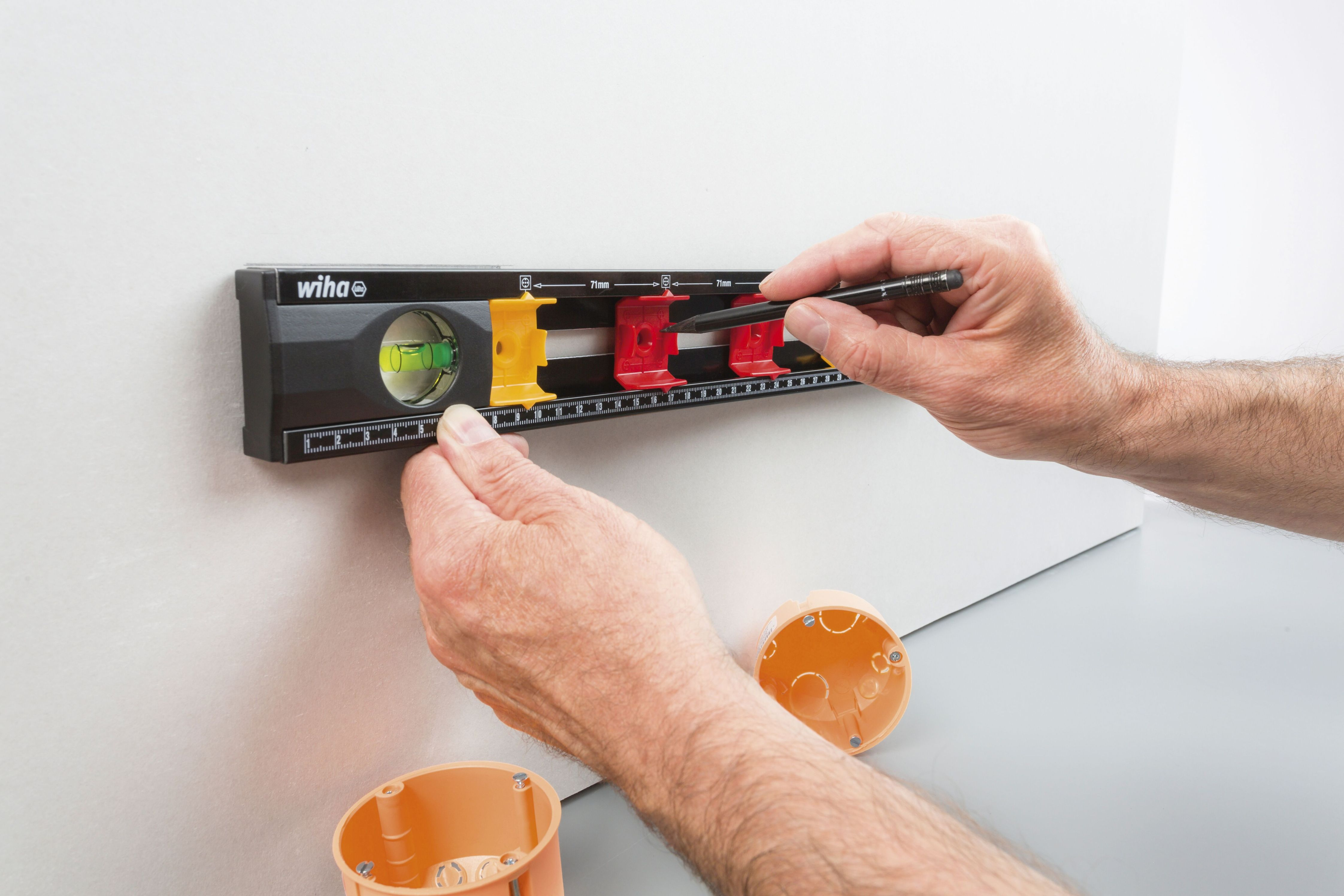 Person benutzt eine Wasserwaage an einer Wand. Die Wasserwaage ist schwarz mit gelben und roten Markierungen. Orange Dose.