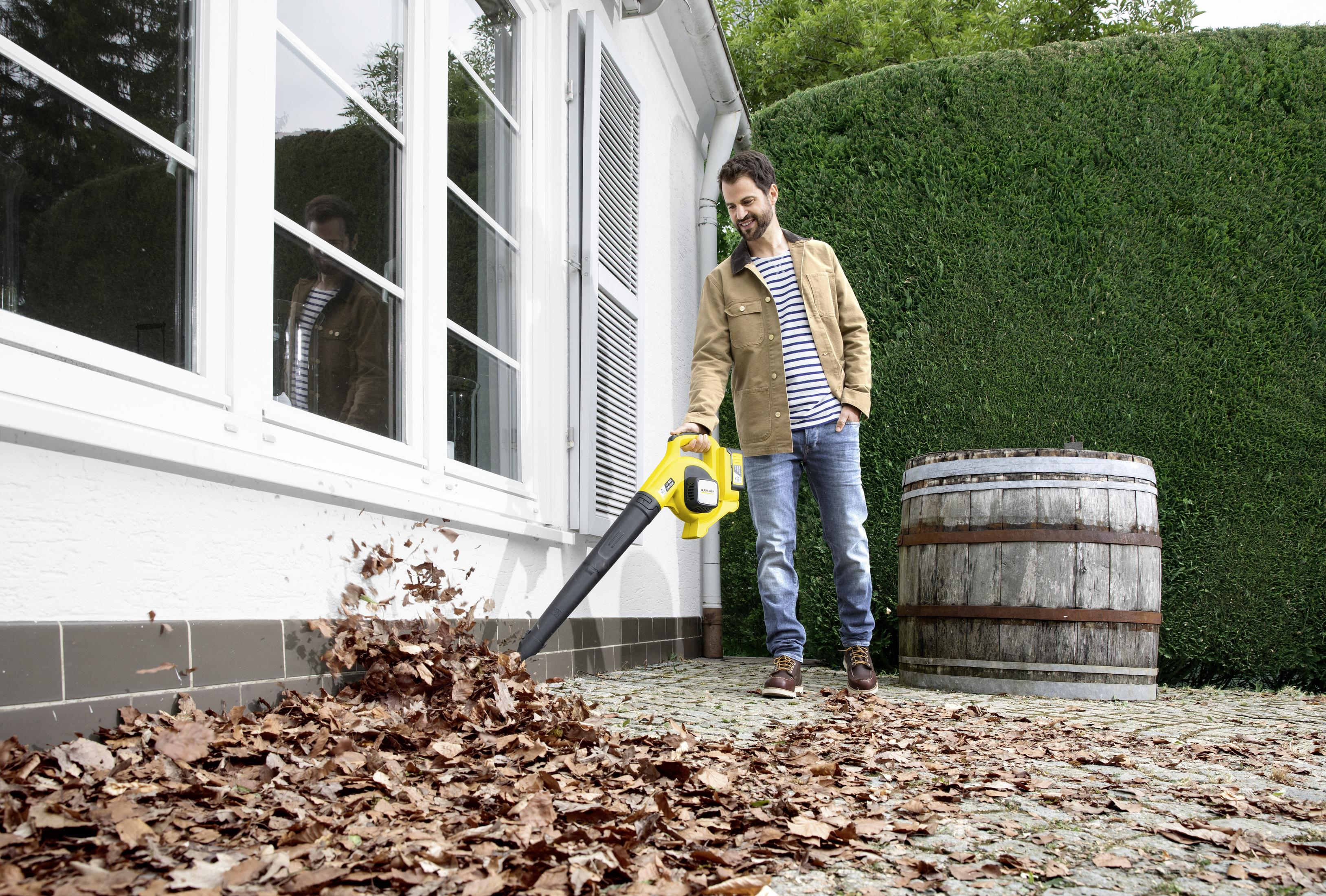 Mężczyzna używa żółtej dmuchawy do liści w pobliżu białego budynku z żywopłotem i drewnianą beczką.