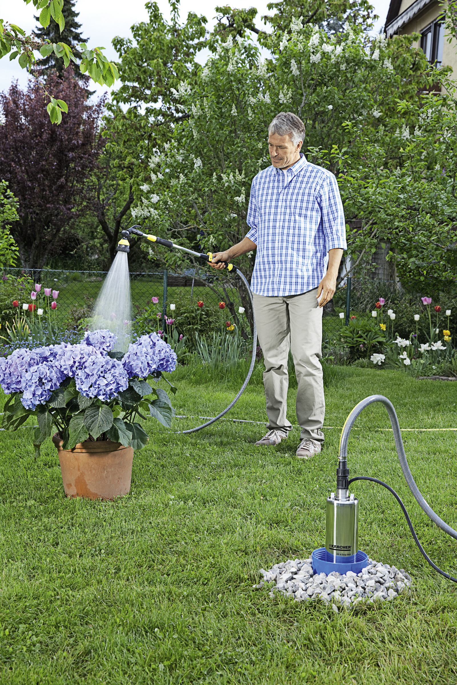 Hombre regando flores azules en maceta con manguera. Césped verde, árboles y bomba en el fondo.