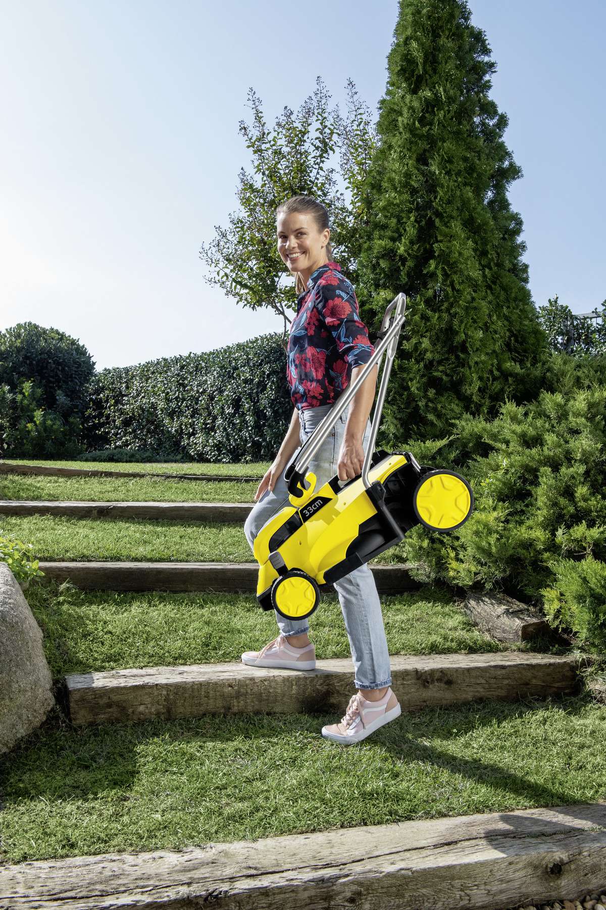Eine Frau trägt einen gelben Rasenmäher eine Holztreppe in einem Garten hinunter.