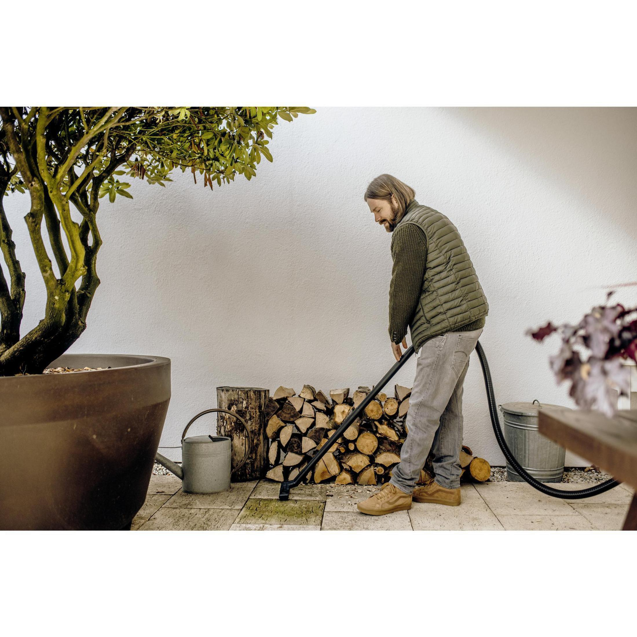 Mężczyzna używa odkurzacza na zewnątrz. Stoi w pobliżu stosu drewna i konewki.