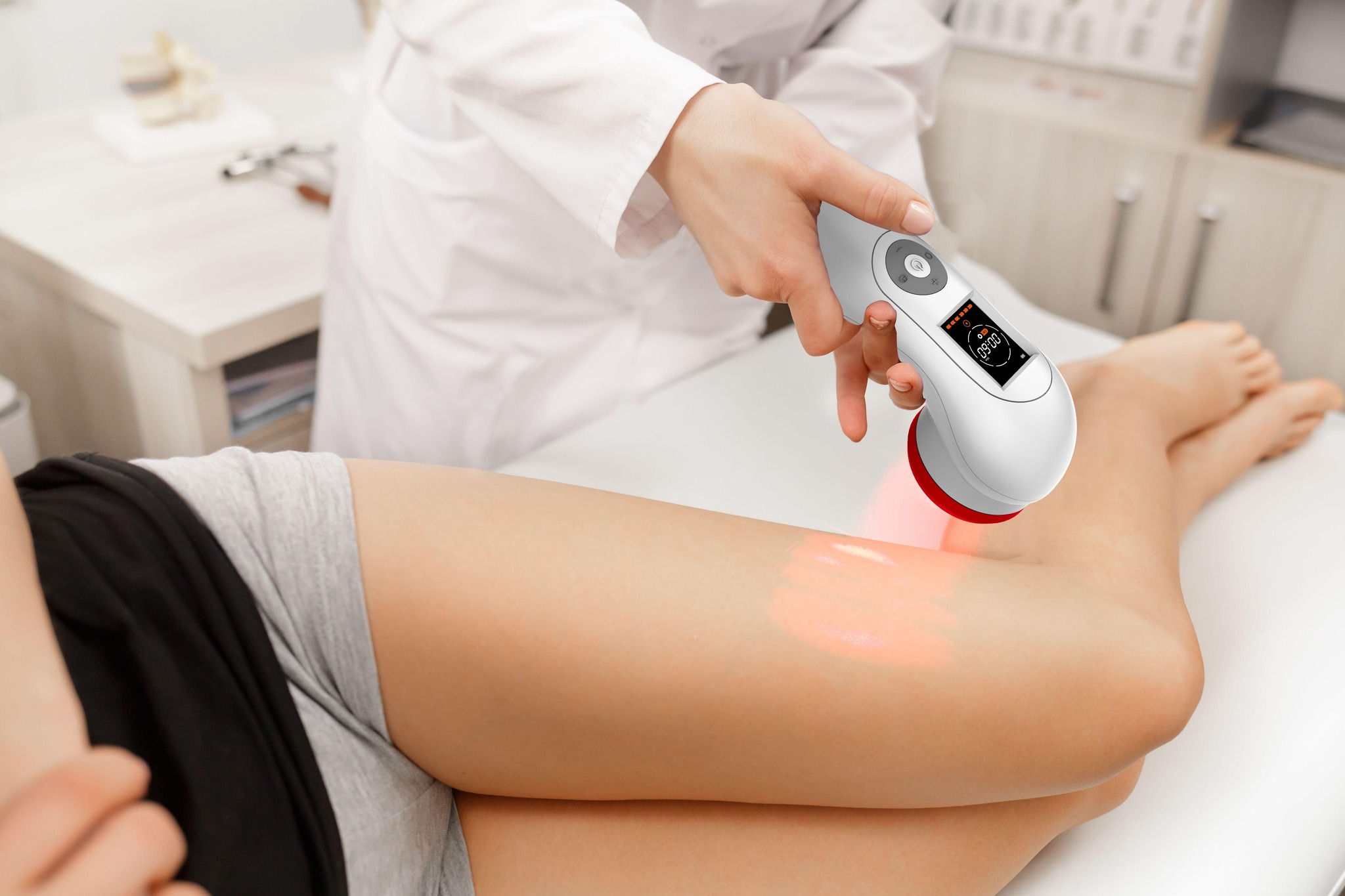 Person benutzt ein Rotlichttherapiegerät am Bein. Weißer Raum. Gerät ist weiß und grau.