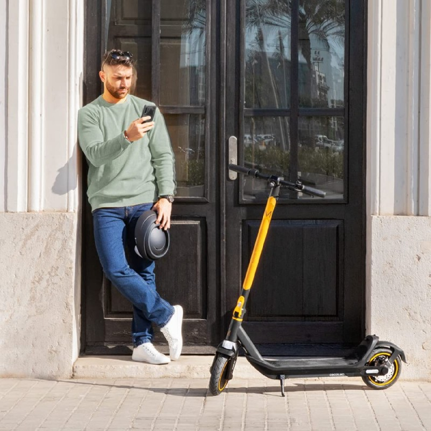 Uomo in piedi vicino a una porta, guarda il telefono, con uno scooter alla sua destra. Tiene un casco.