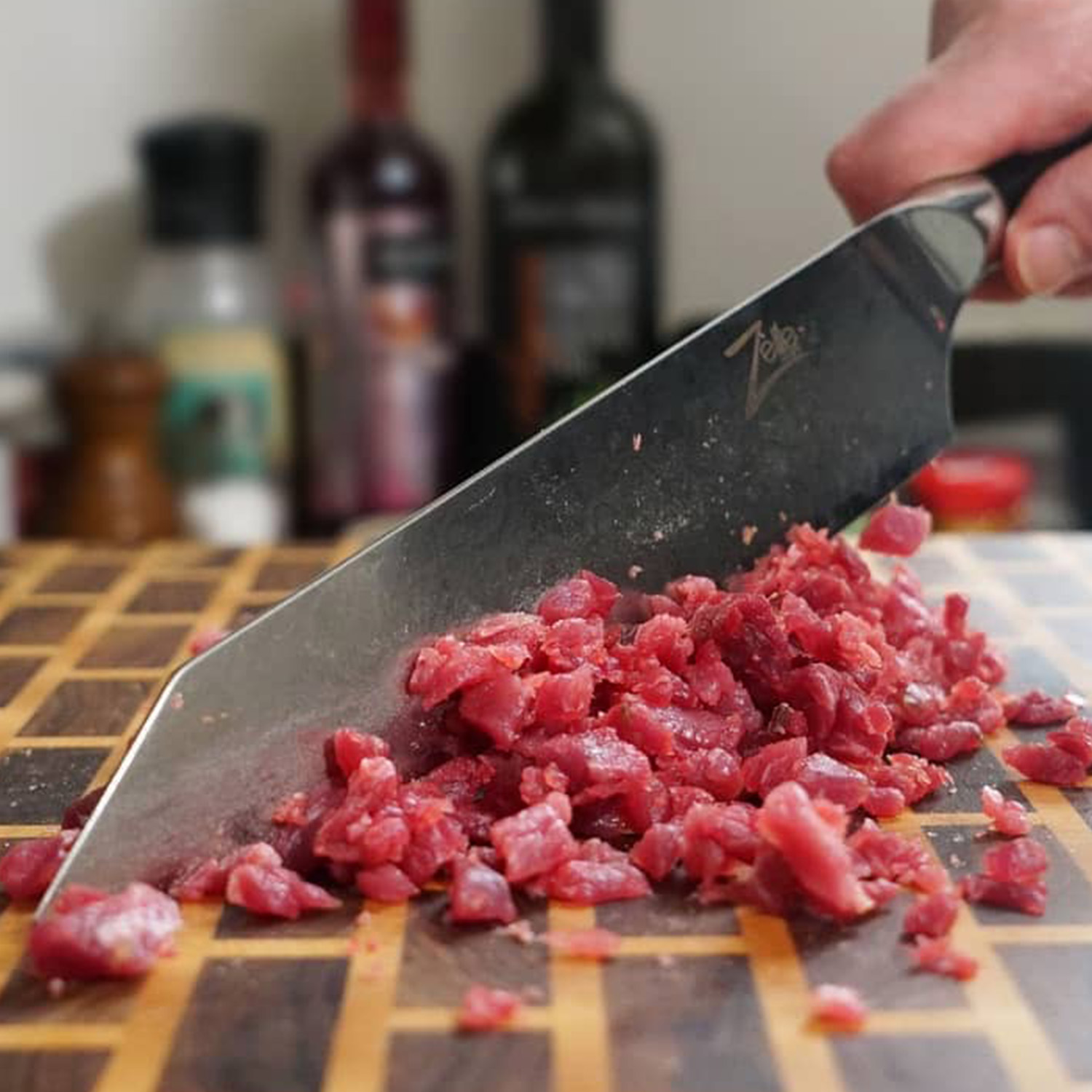 Ein Messer schneidet gewürfeltes Fleisch auf einem hölzernen Schneidebrett. Das Fleisch ist rot und das Messer silberfarben.