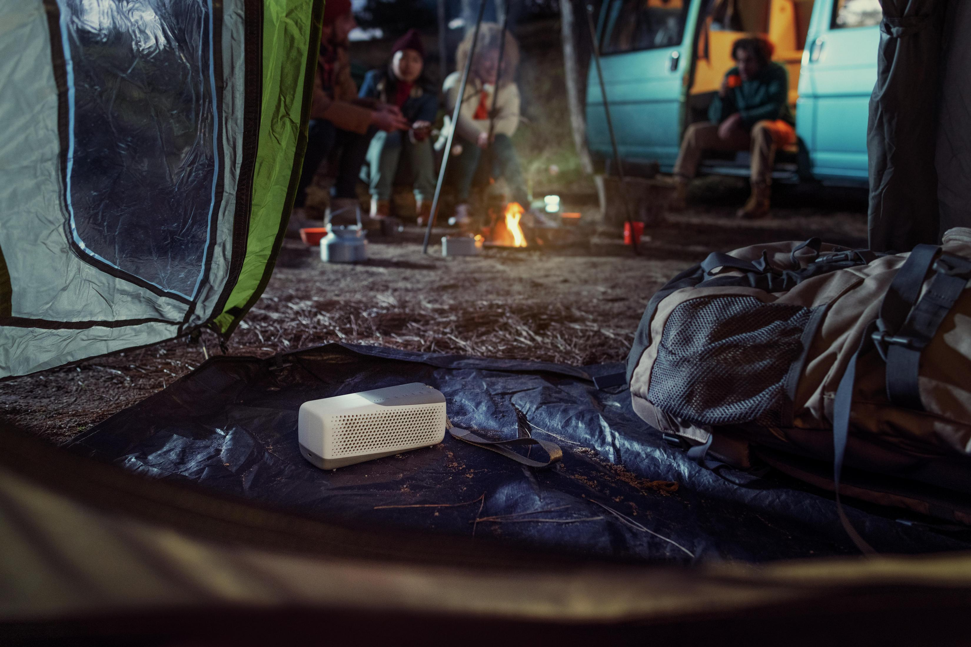 Głośnik na ciemnej powierzchni w namiocie, z ludźmi i ogniskiem w tle.