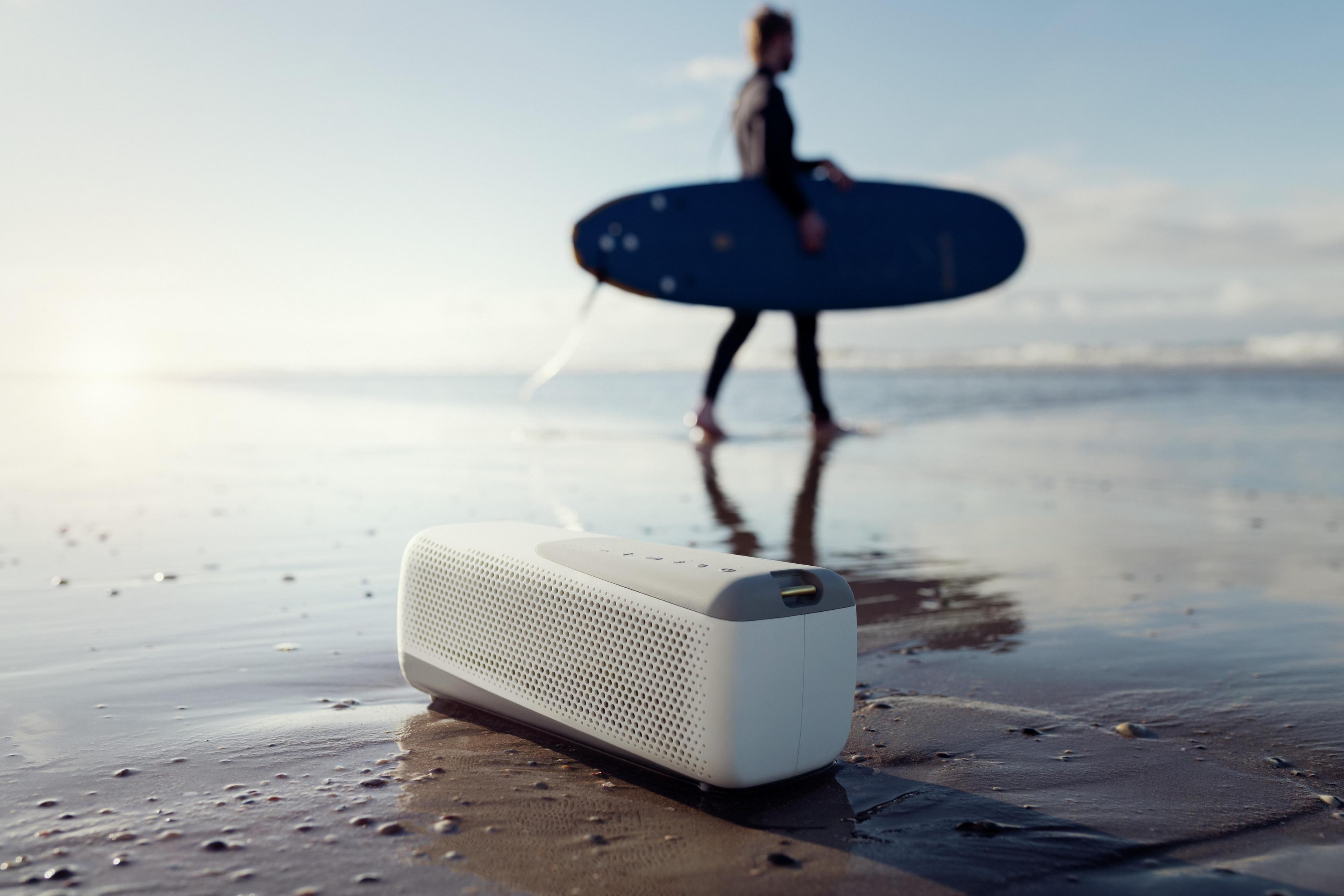 Biały głośnik na plaży, osoba z deską surfingową w tle.