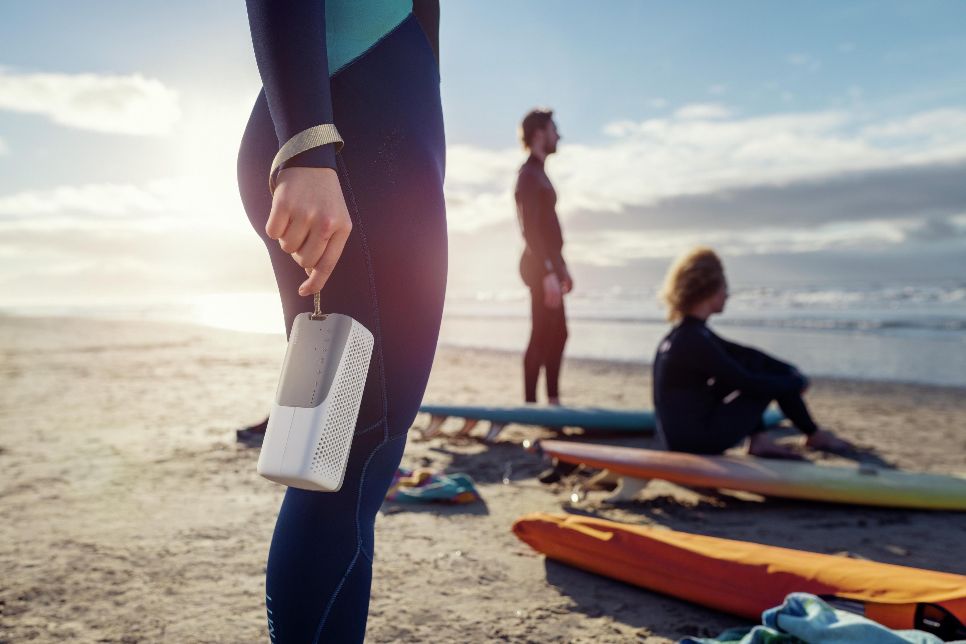 Osoba trzymająca biały głośnik na plaży. Surferzy w tle. Błękitne niebo.