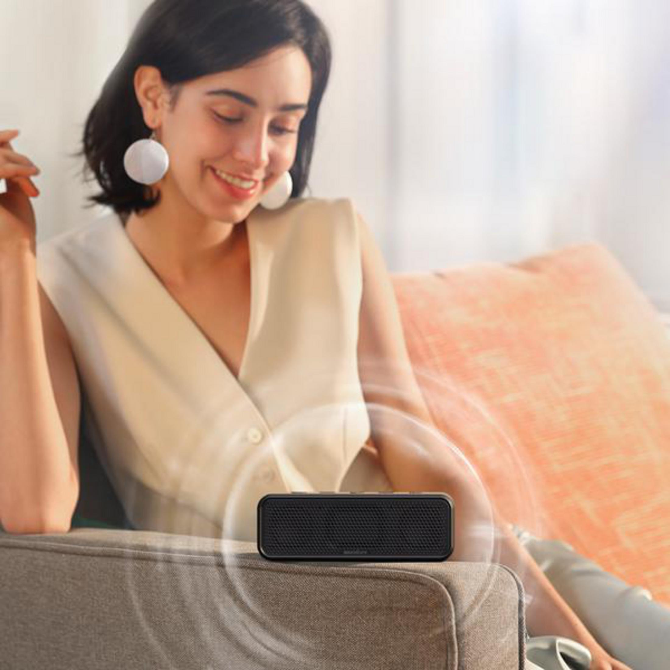 Kobieta siedzi, uśmiecha się, z małym czarnym głośnikiem na kanapie emitującym fale dźwiękowe.