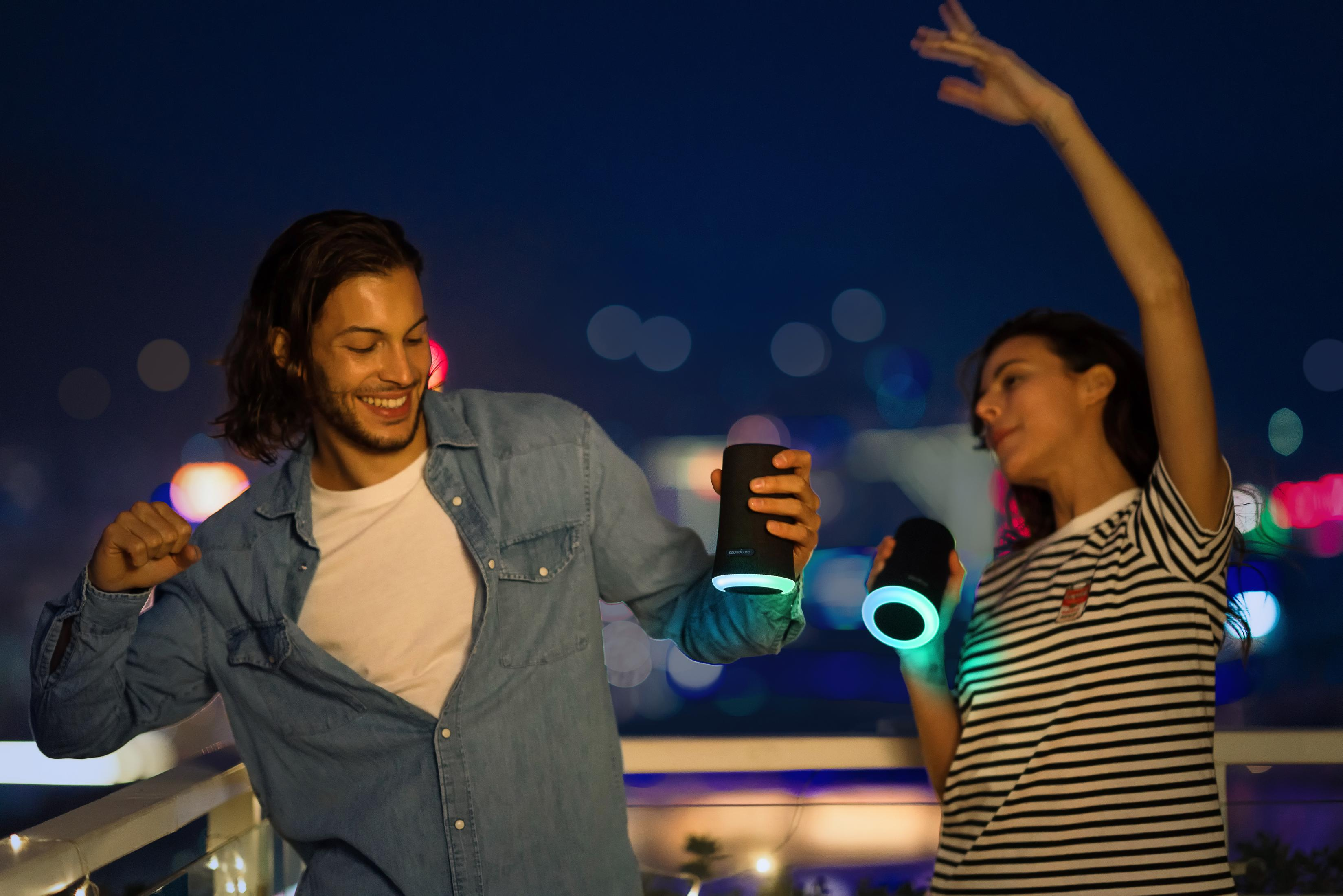 Dwie osoby tańczące z głośnikami w nocy. Światła miasta w tle.