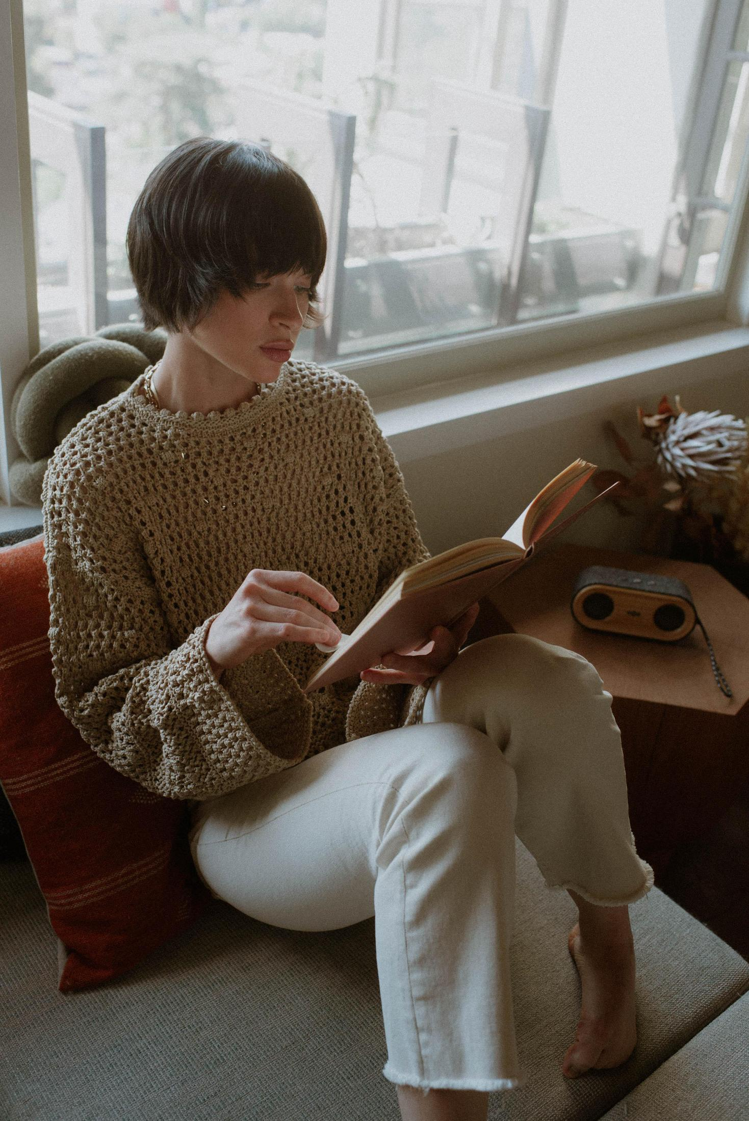 Kobieta czyta książkę. Kobieta siedzi, czyta książkę, w pobliżu okna, z małym głośnikiem.