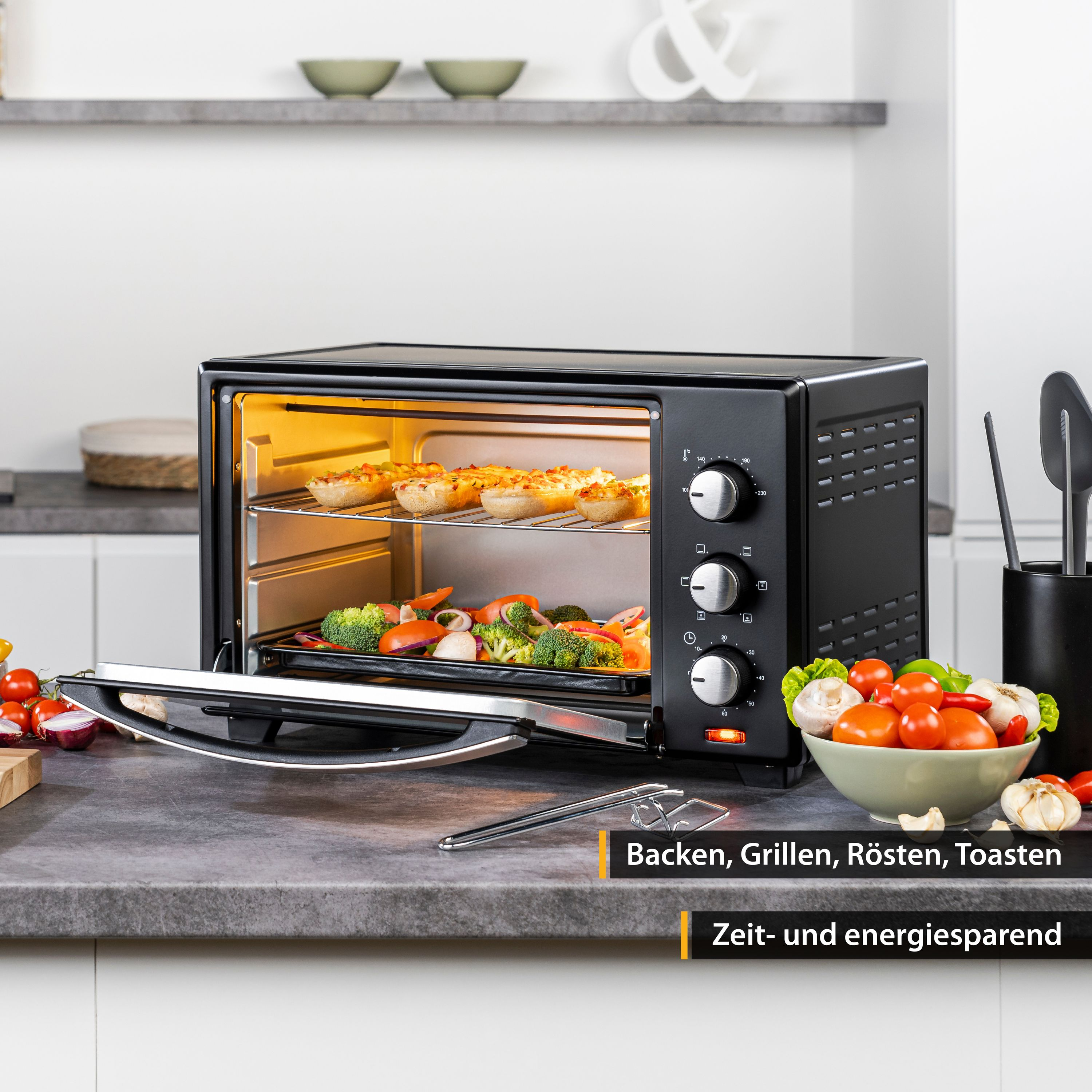Schwarzer Ofen mit Essen darin, Tomaten und Gemüse davor, Text unten.