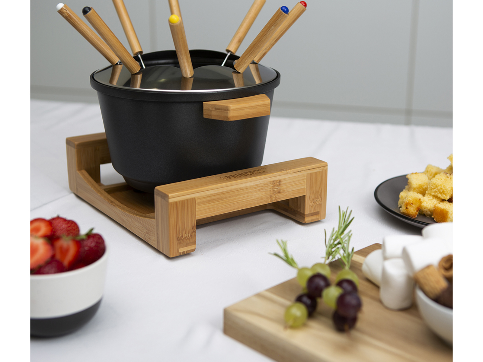 Fondue-Set mit schwarzem Topf, Bambusständer, verschiedenen Spießen und Speisen auf weißer Oberfläche.