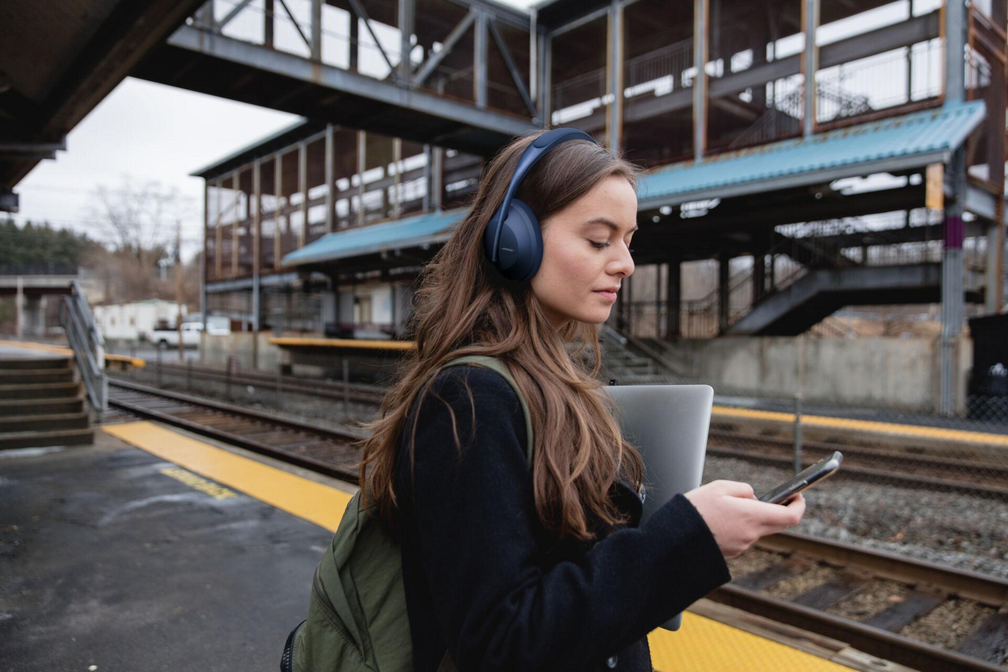 Eine Frau mit Kopfhörern auf einem Bahnsteig, die ein Telefon benutzt und einen Laptop hält.