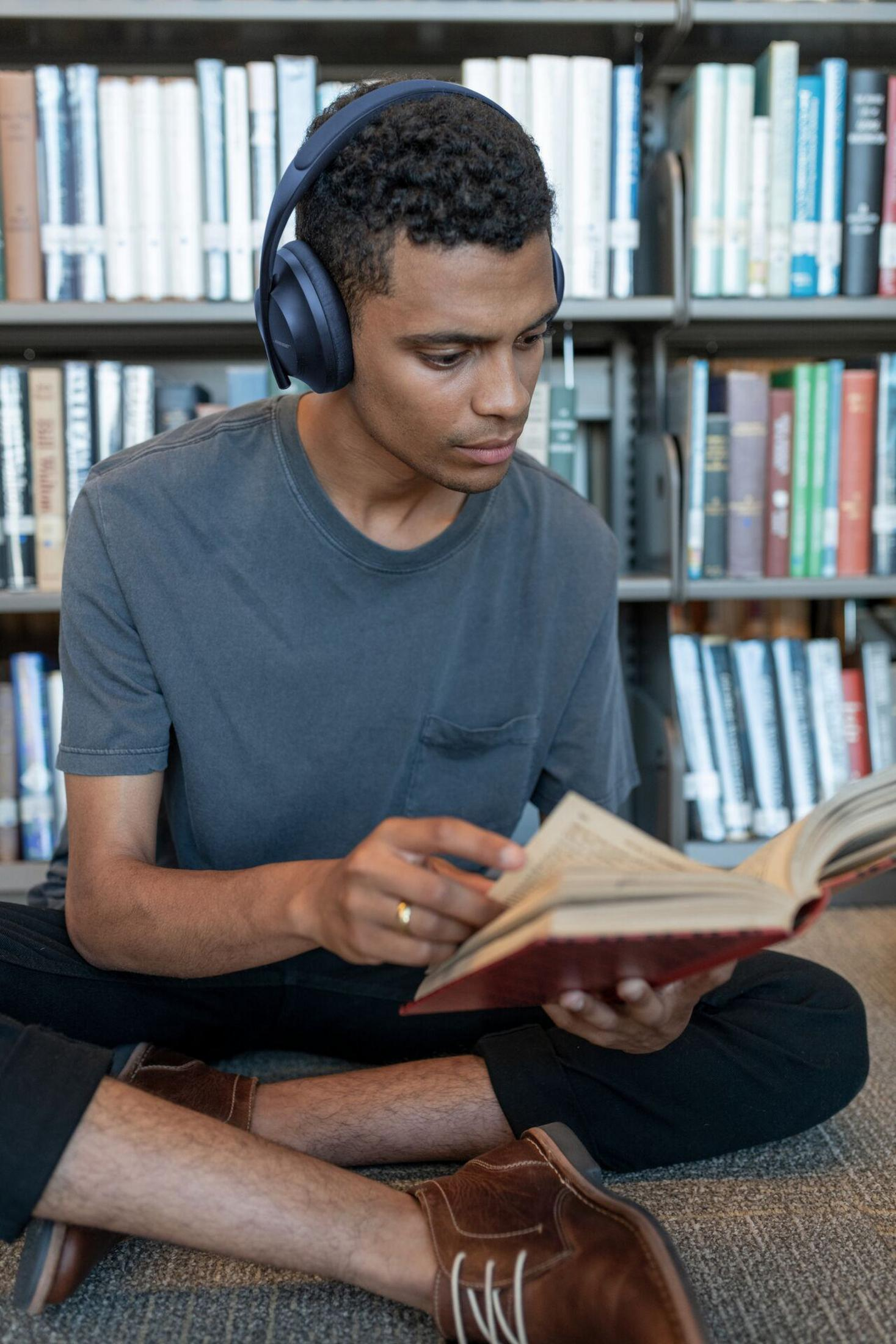 Ein junger Mann mit blauen Kopfhörern liest ein Buch in einer Bibliothek, sitzt auf dem Boden.
