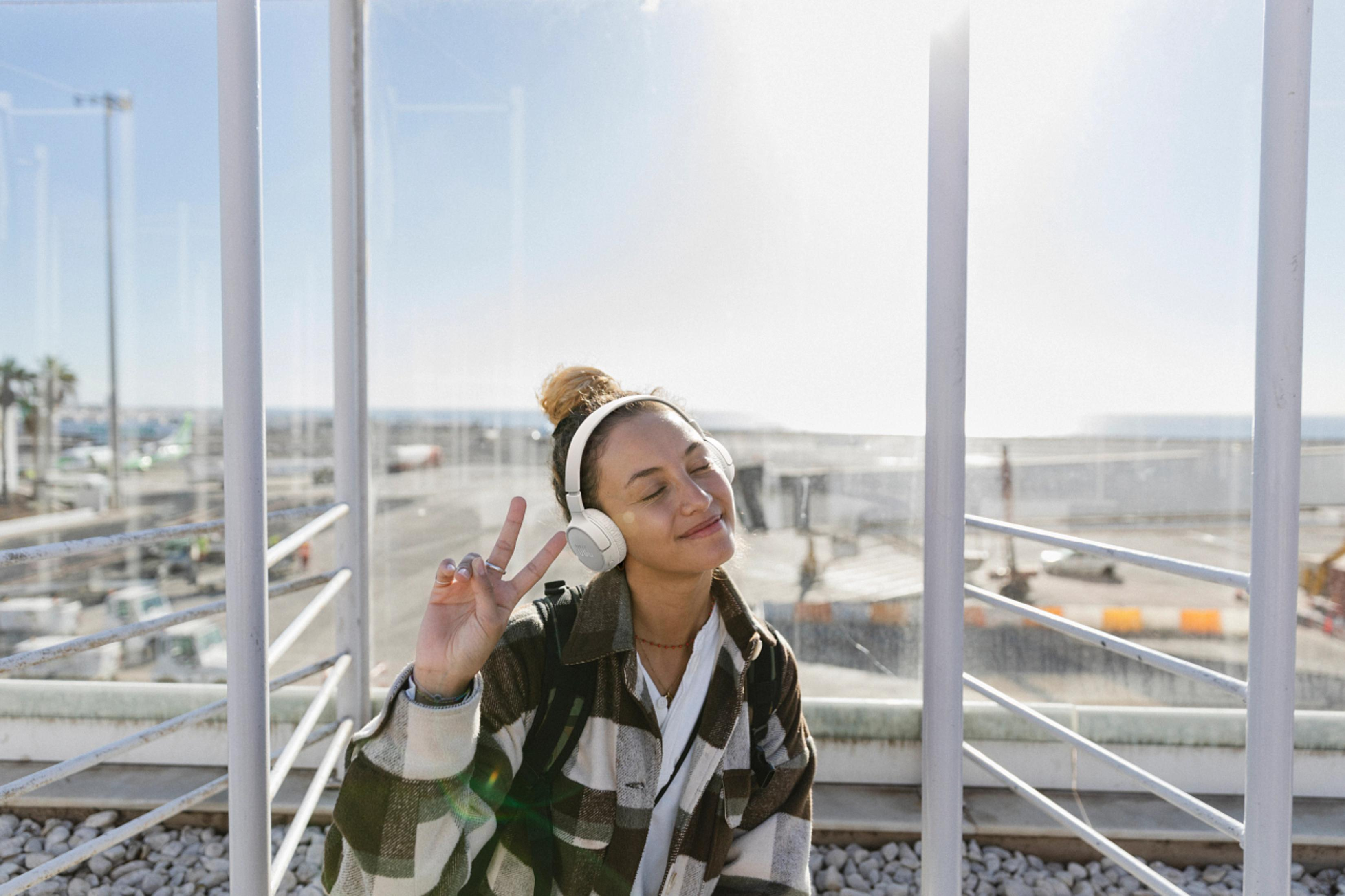 Eine Frau mit Kopfhörern macht ein Friedenszeichen vor einem klaren Fenster mit sonnigem Hintergrund.