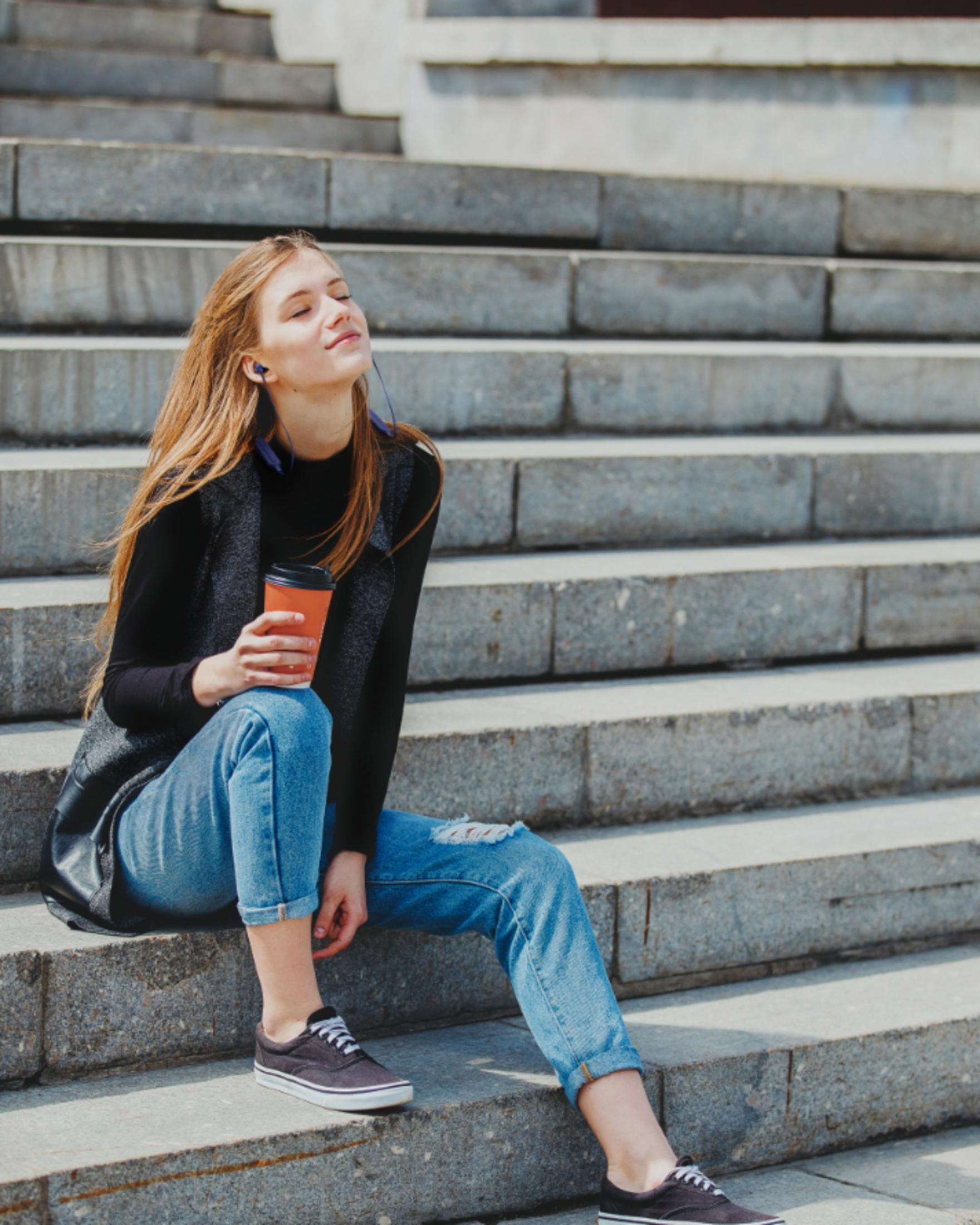 Kobieta siedząca na schodach, trzymająca kubek kawy i słuchająca muzyki.