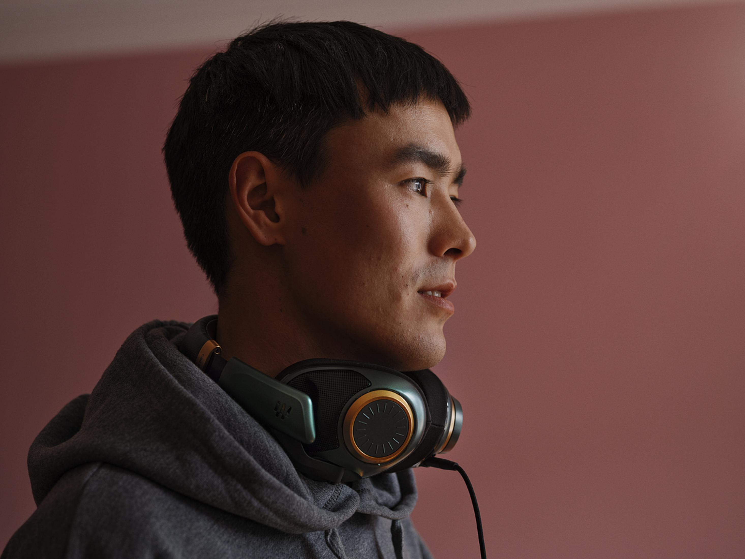 Mężczyzna w szarej bluzie z kapturem i zielono-złotych słuchawkach, patrzący w prawo.