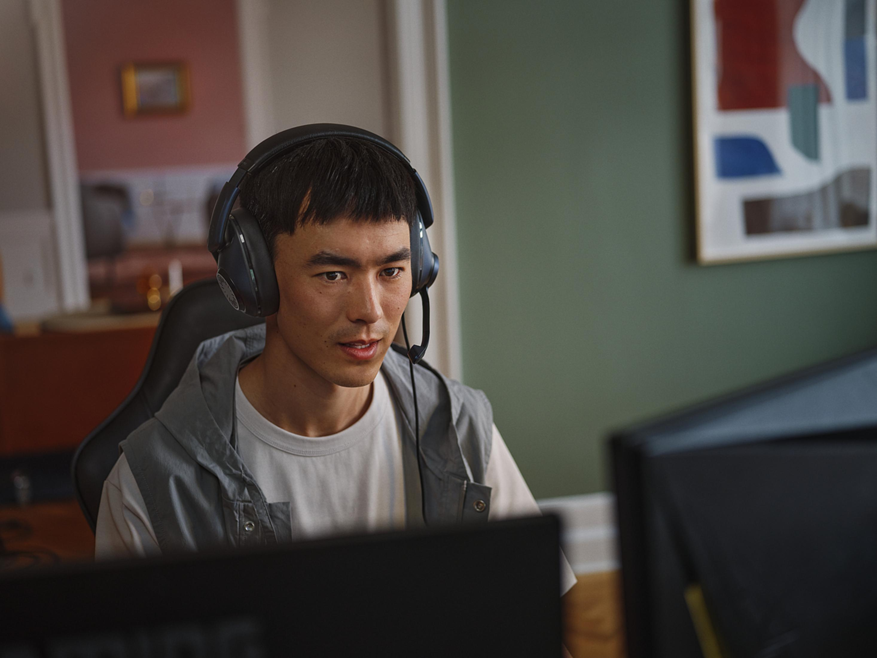 Mężczyzna w zestawie słuchawkowym, gra w pokoju. Patrzy na monitor. Tło: zielona ściana, obraz.