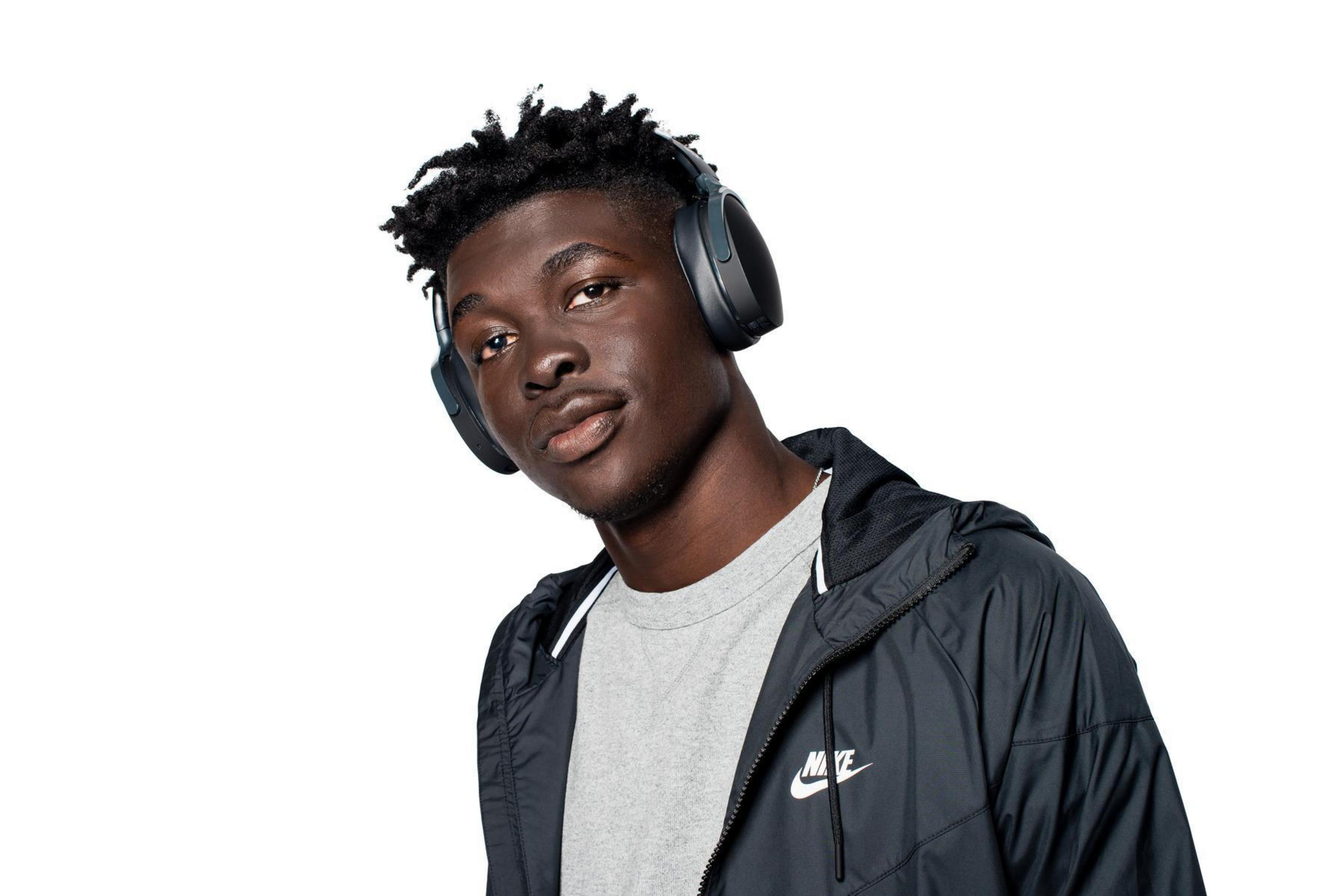 Młody czarny mężczyzna w słuchawkach, szarej koszulce i czarnej kurtce. Patrzy w kamerę.