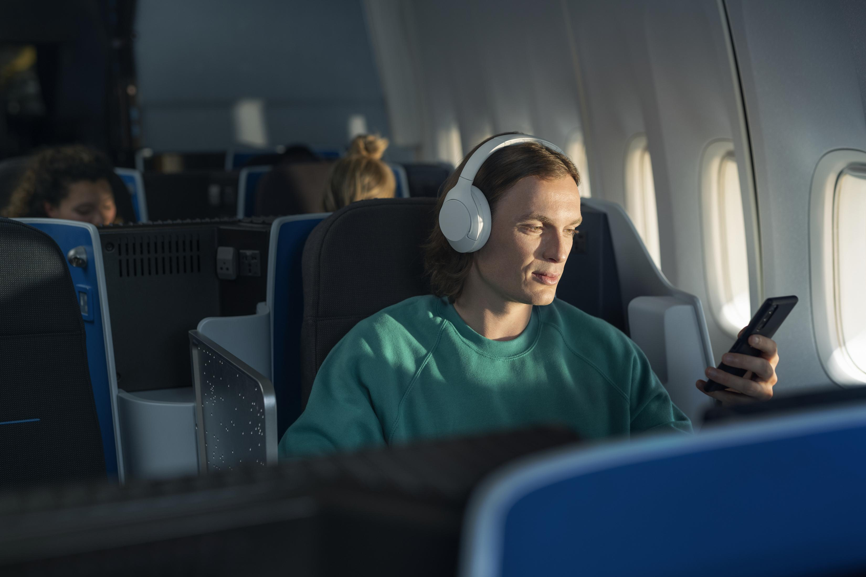 Ein Mann im Flugzeug mit weißen Kopfhörern, der auf ein Telefon schaut.