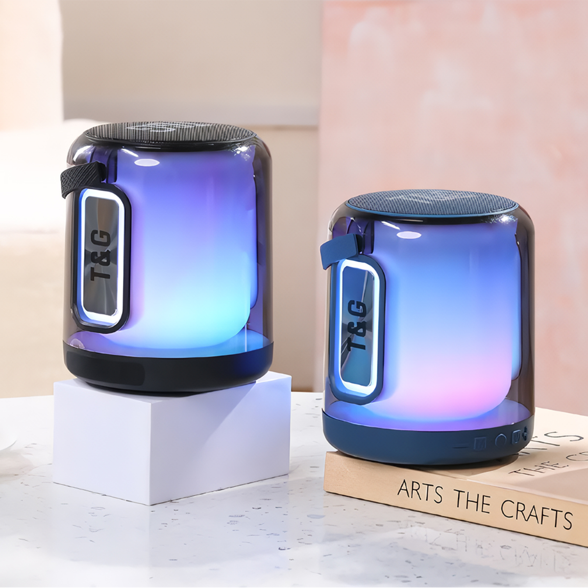 Dos altavoces cilíndricos con luces LED, sobre cubos blancos. Libro y fondo rosa.