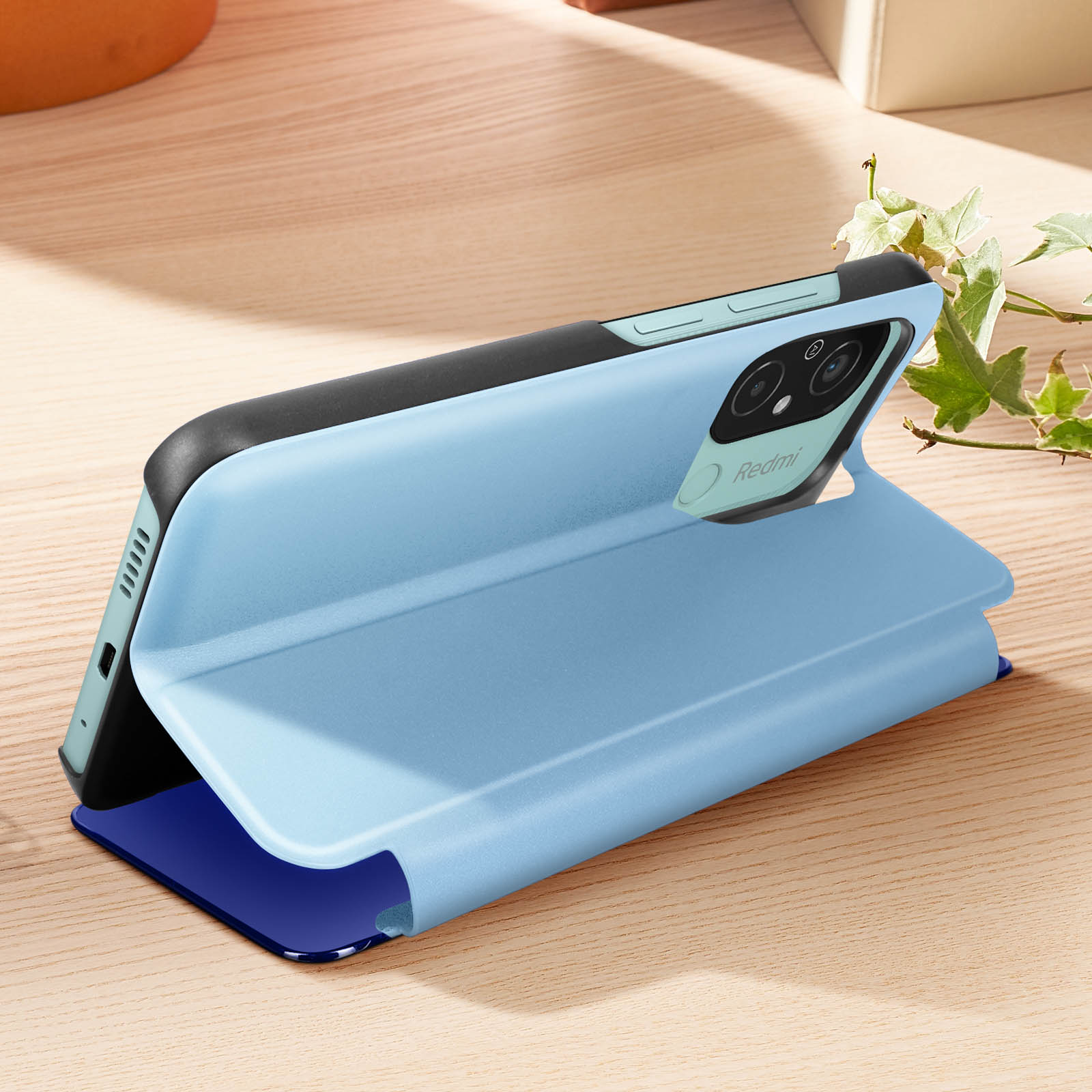 Custodia blu per telefono aperta, che mostra un telefono con cornice nera, su legno e una pianta.