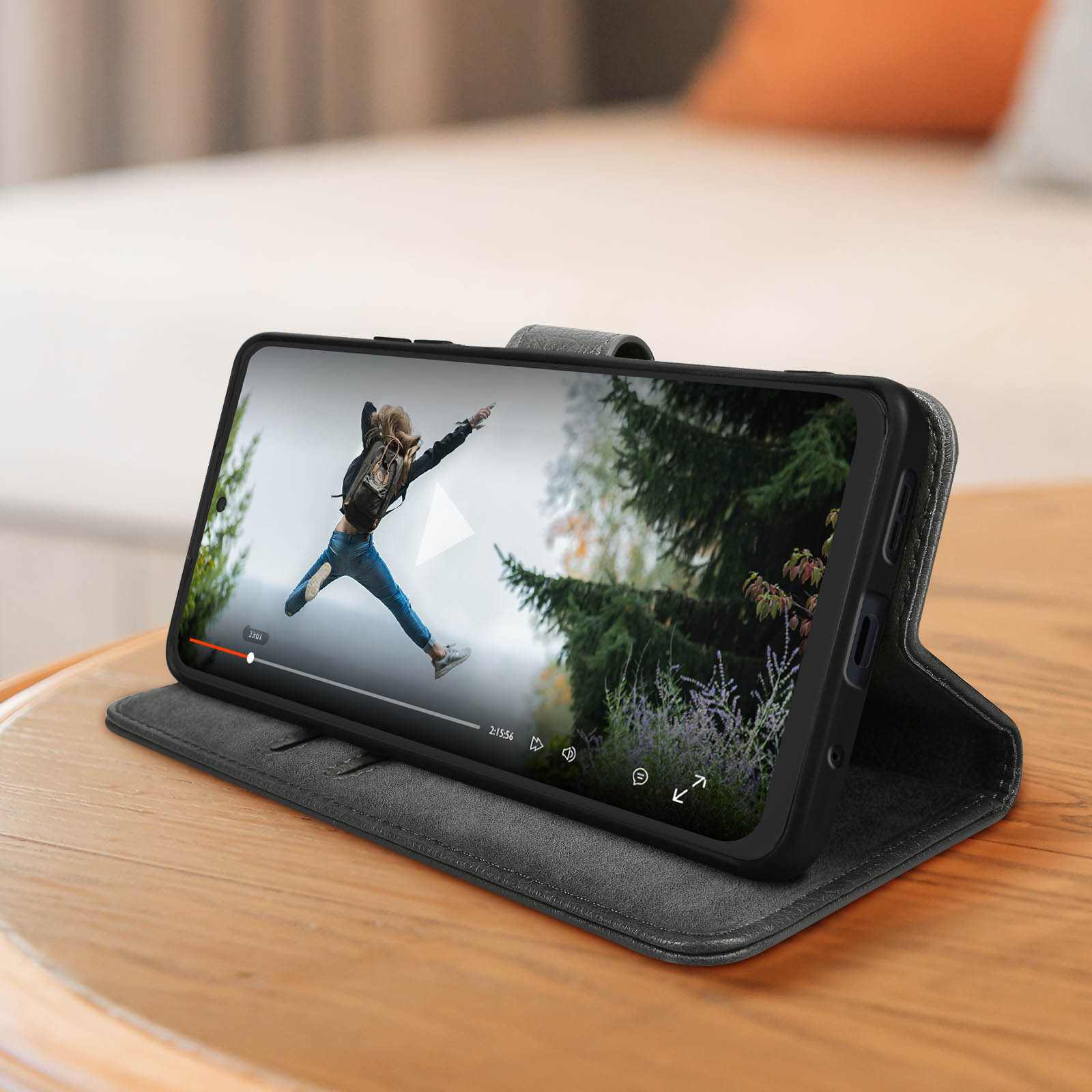 Smartphone in custodia su un tavolo di legno. Lo schermo mostra una persona che salta. Lo sfondo è sfocato.