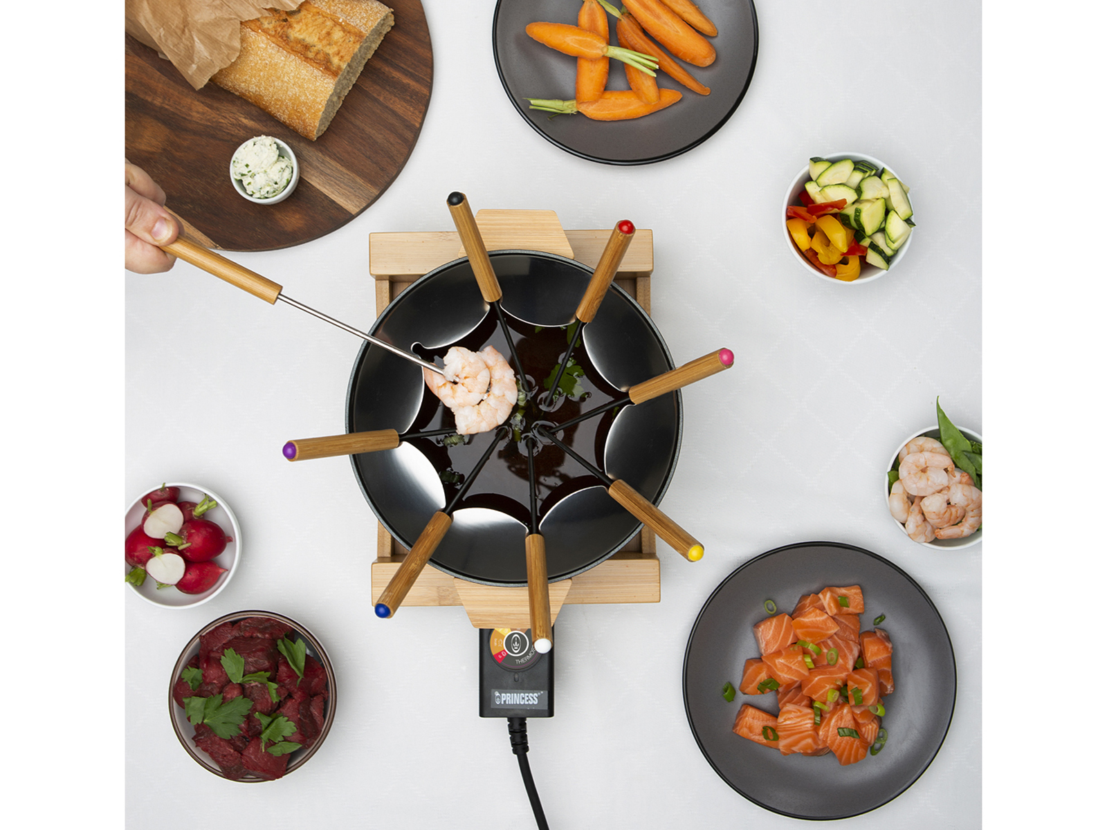 Garnek do fondue ze składnikami dookoła: warzywa, mięso, chleb. Ręka używa widelca.