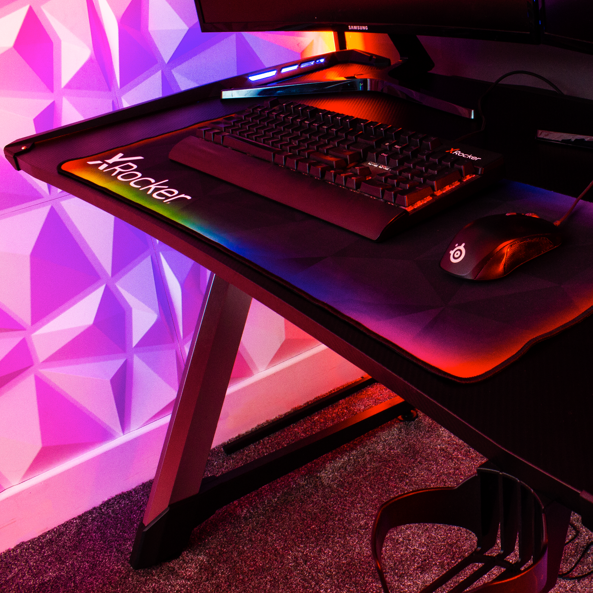 Ein Gaming-Schreibtisch mit Tastatur, Maus und Mauspad mit farbiger Beleuchtung an einer weißen Wand.