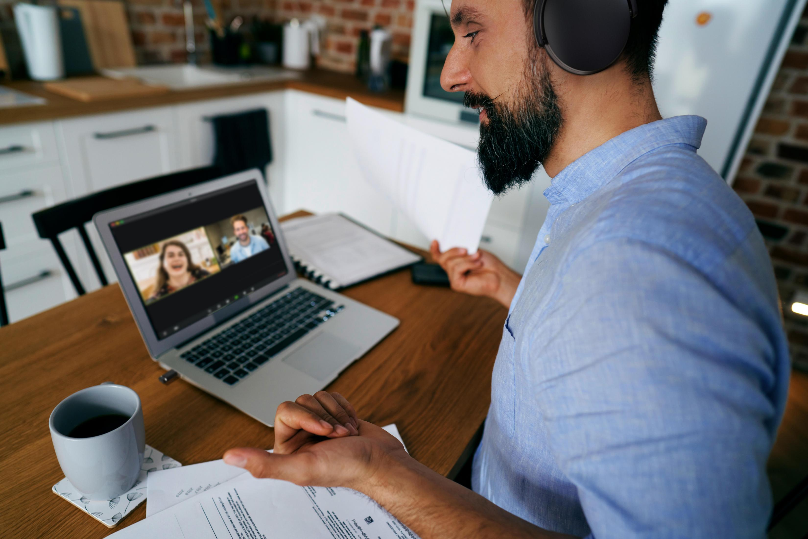 Uomo in camicia blu in videochiamata con due persone su un laptop, con documenti e una tazza di caffè.