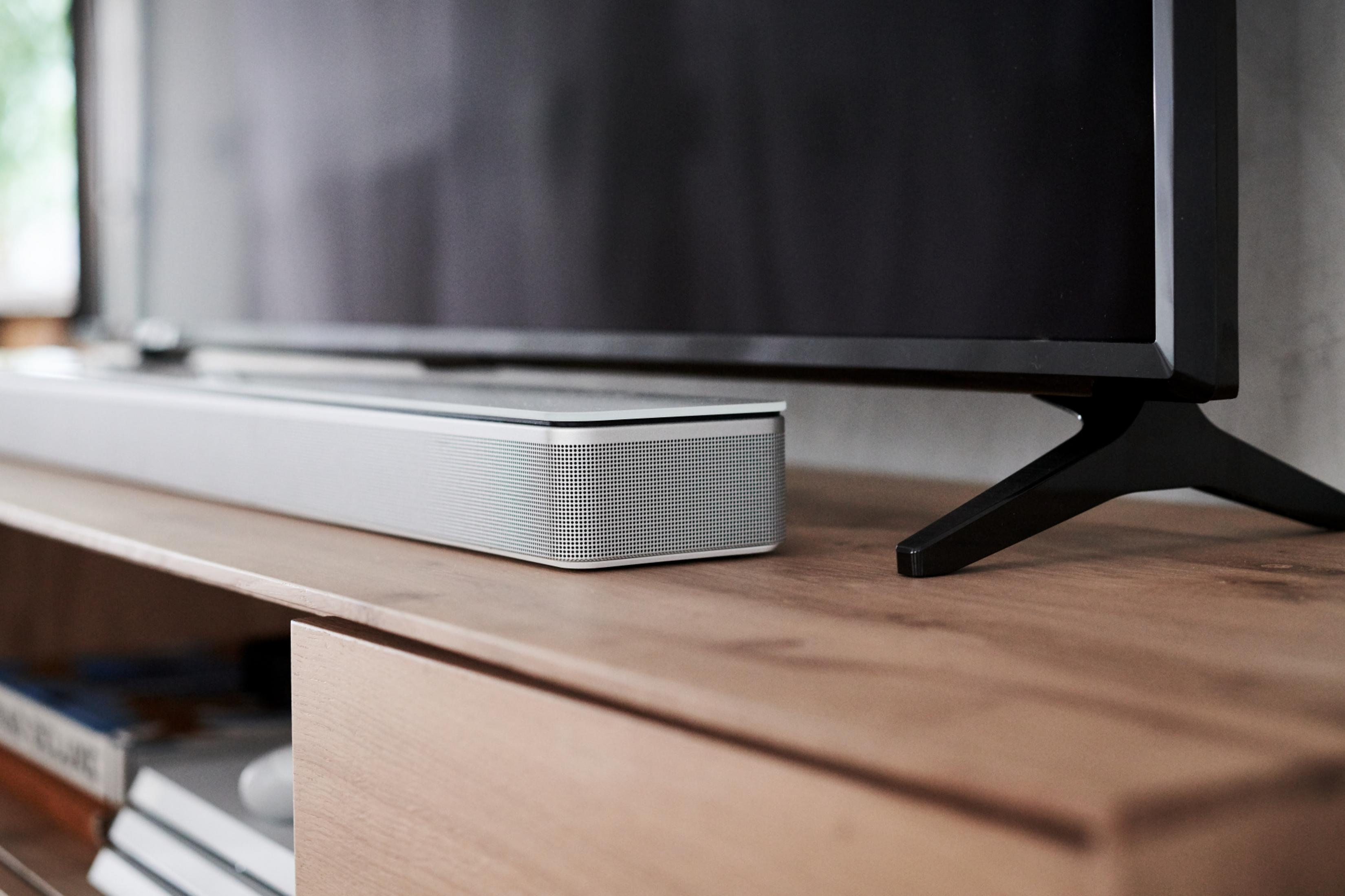 Silberne Soundbar auf Holzoberfläche vor einem schwarzen Fernseher auf einem Ständer. Bücher sind sichtbar.