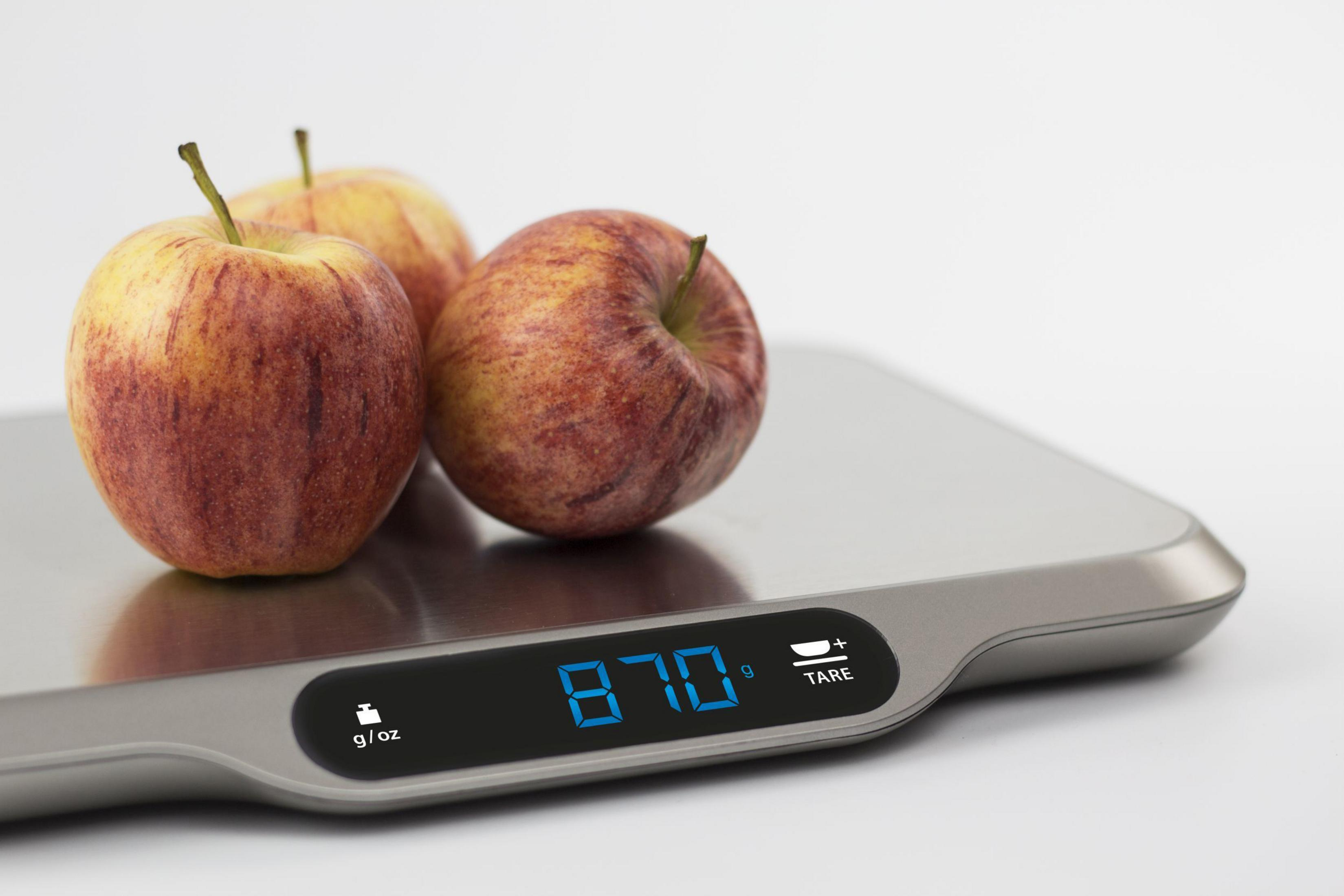 Drei Äpfel auf einer silbernen Waage, Digitalanzeige zeigt 870 g/oz. Weißer Hintergrund.