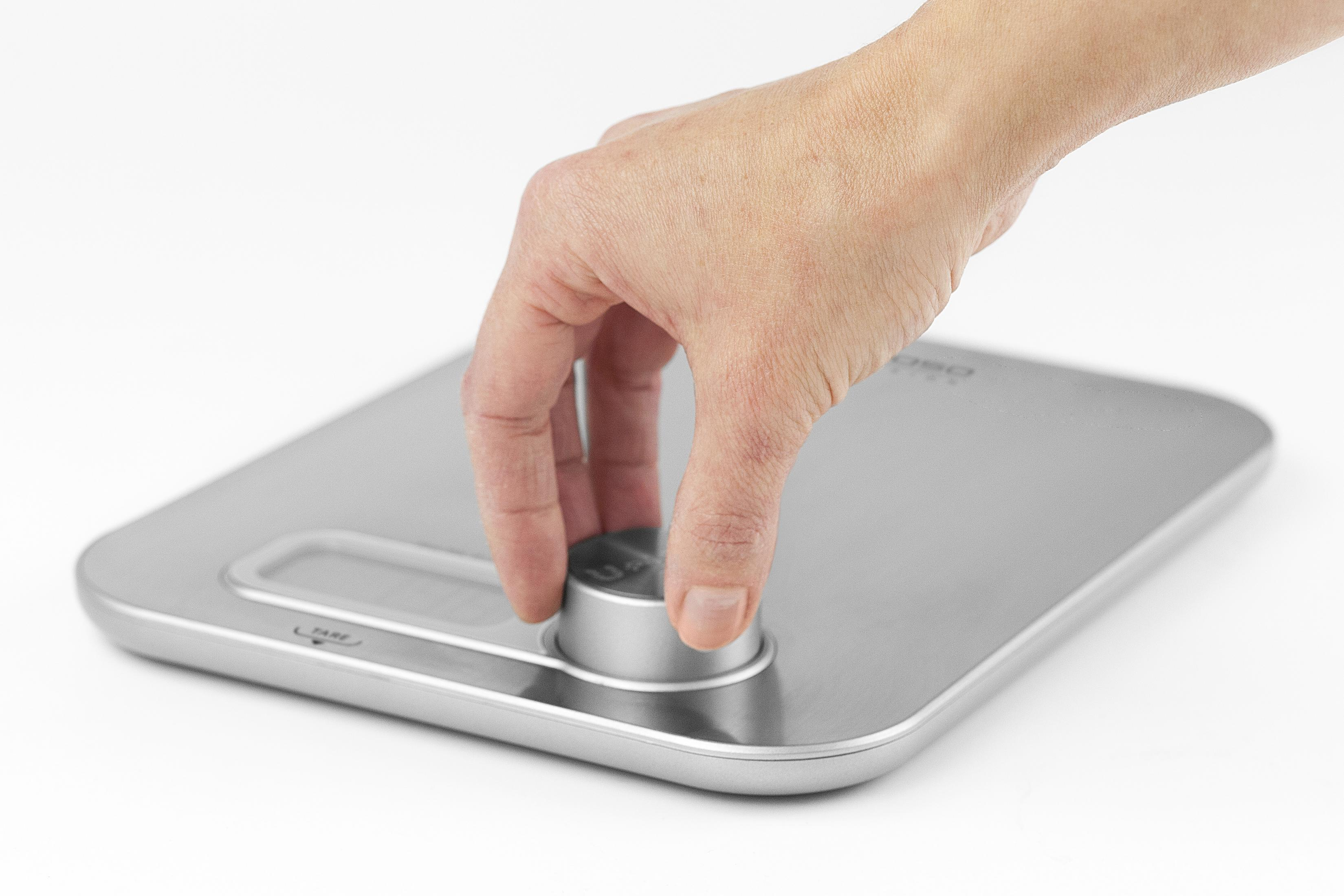 Una mano che ruota un quadrante su una bilancia da cucina argentata. La bilancia è su sfondo bianco.