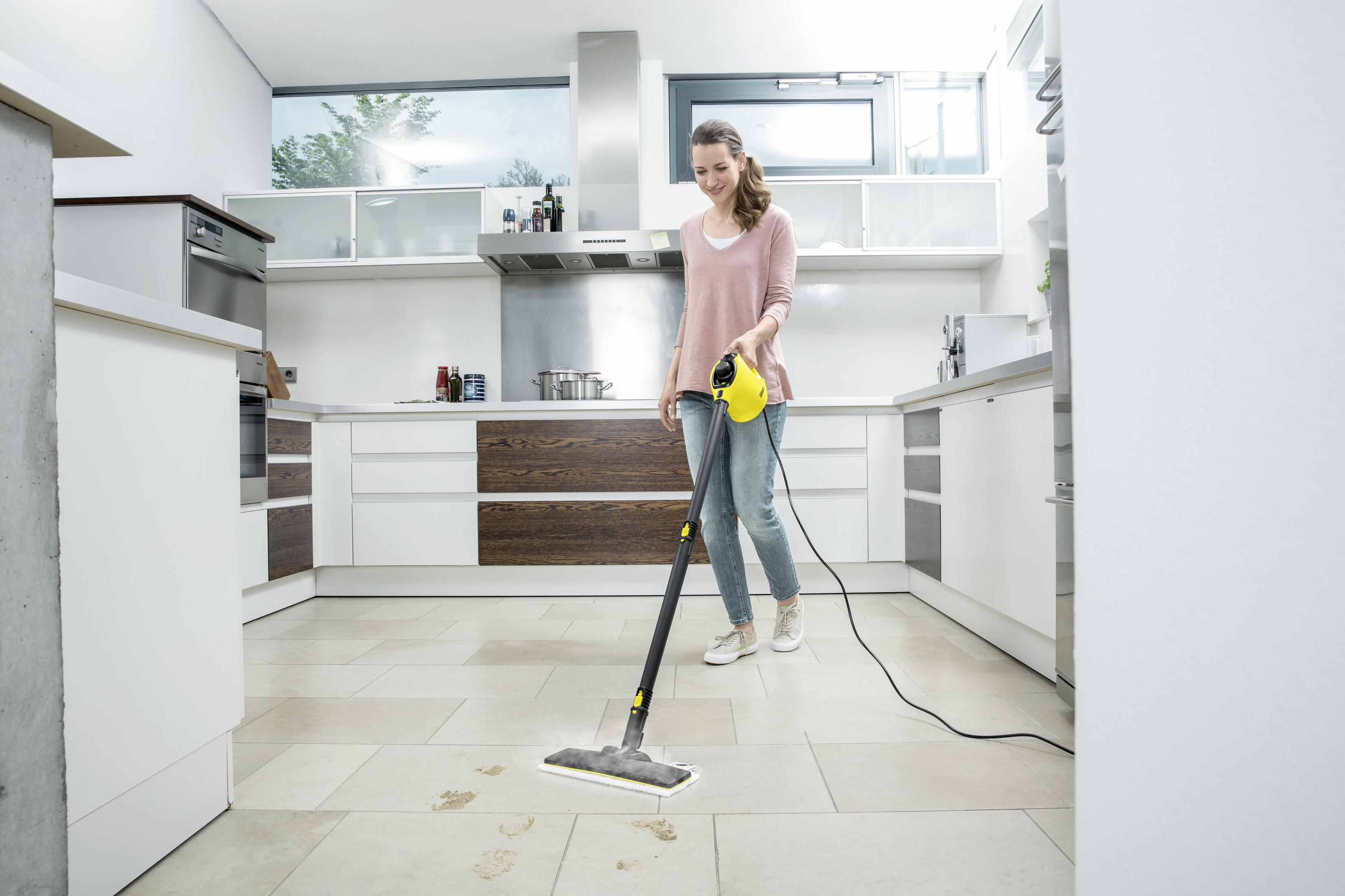 Donna in cucina, pulisce il pavimento piastrellato con una pulitrice a vapore gialla. La cucina ha mobili bianchi.