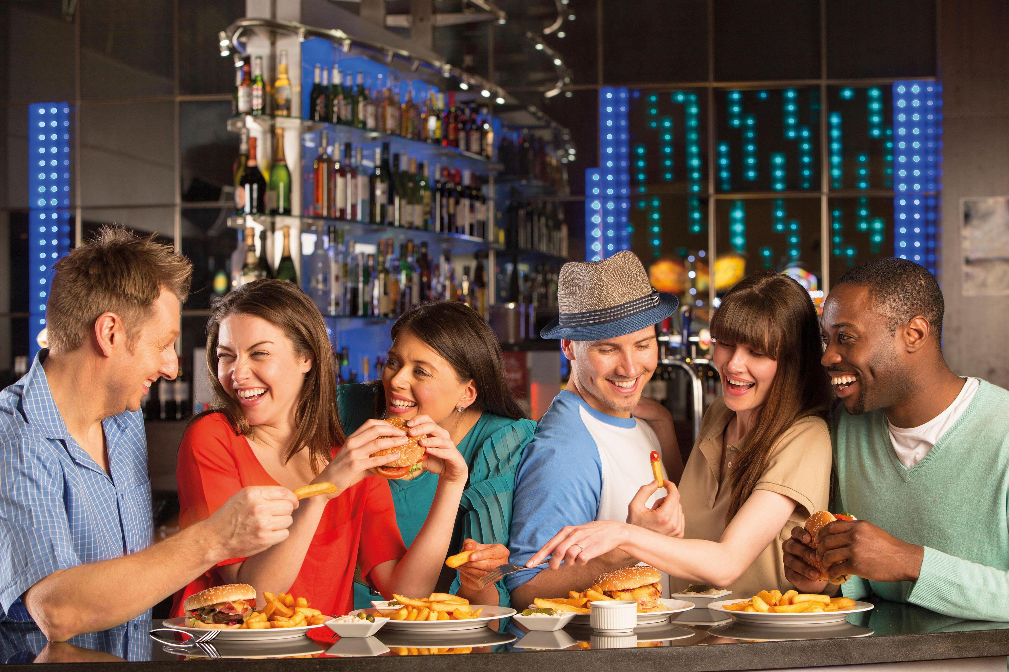 Grupa ludzi jedzących burgery i frytki w barze, śmiejących się. Butelki i światła w tle.