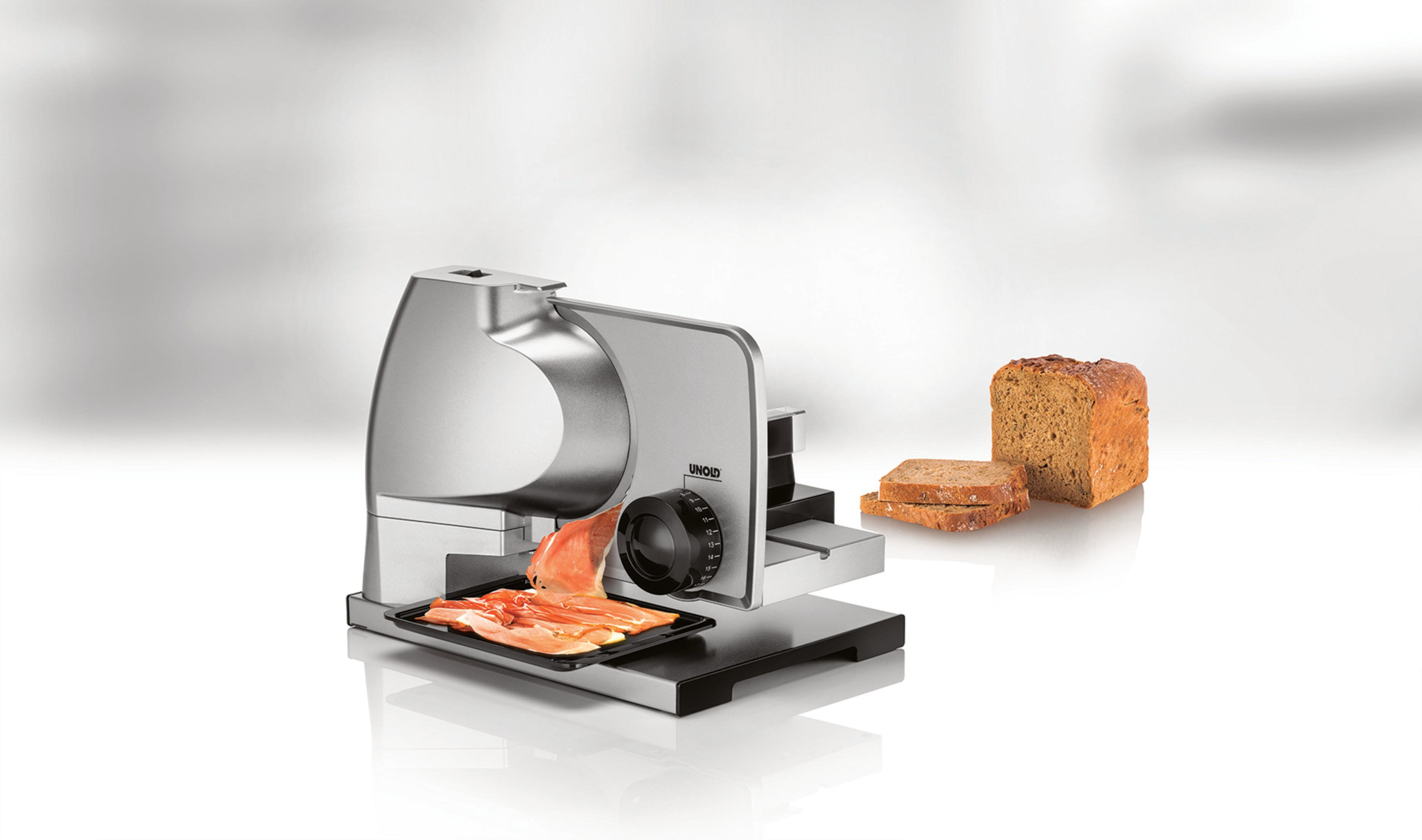 Krajalnica z mięsem i chlebem. Krajalnica jest srebrna, z chlebem po prawej stronie.