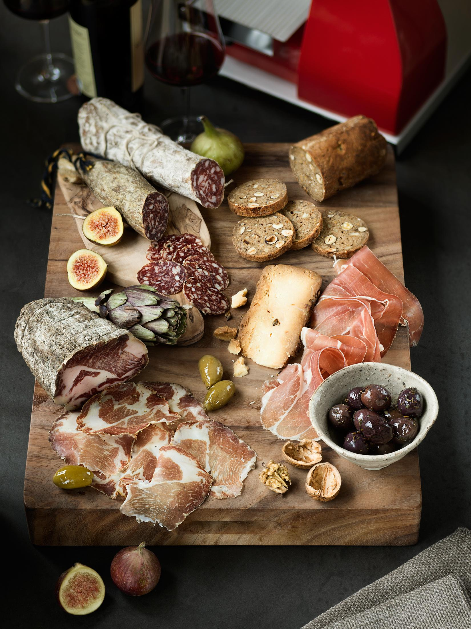 Tagliere in legno con salumi vari: carni, formaggi, olive, fichi, pane e noci.
