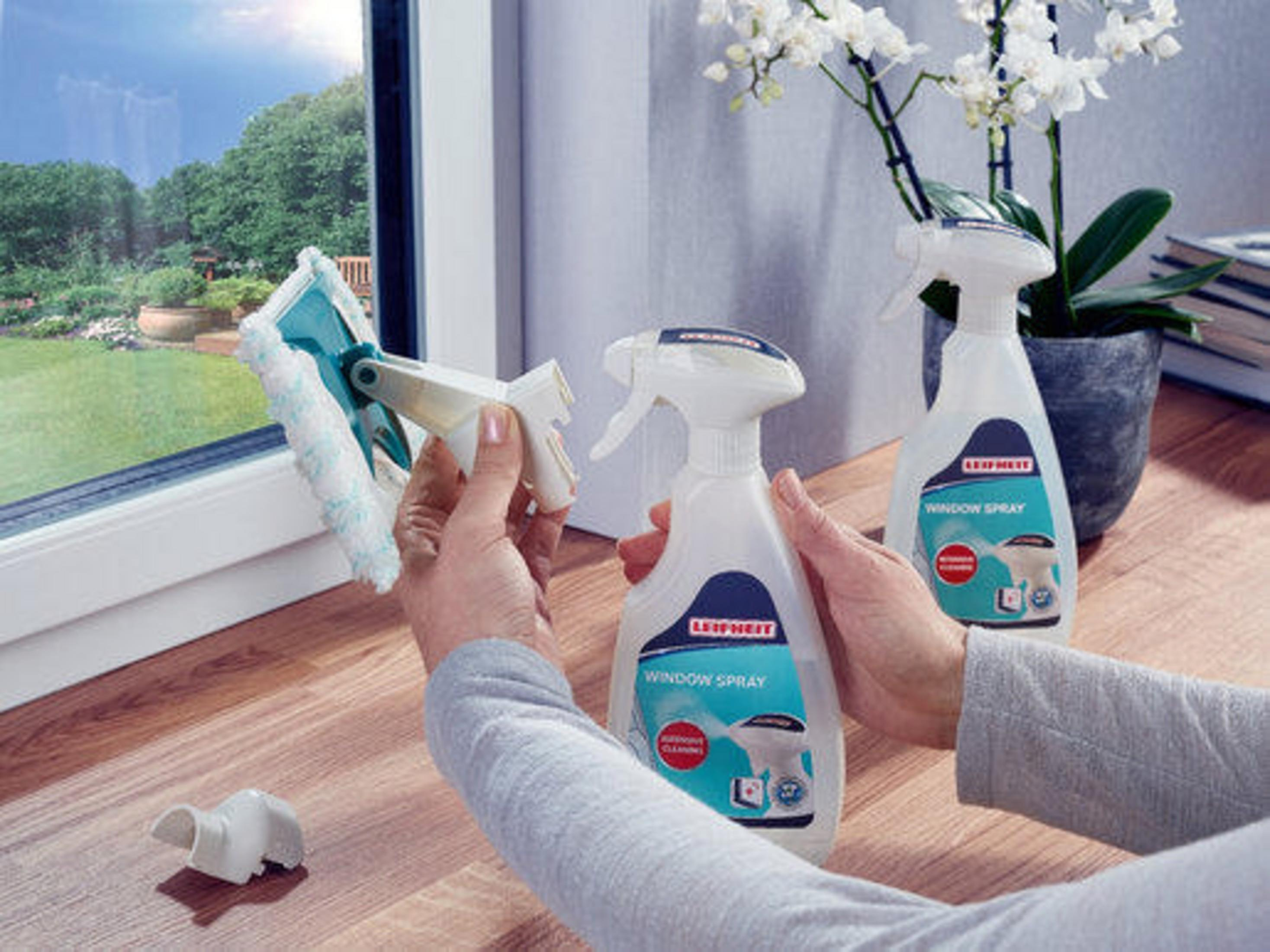 Hände einer Person, die Fensterputzwerkzeuge halten, mit Fenstern und Sprühflaschen im Hintergrund.