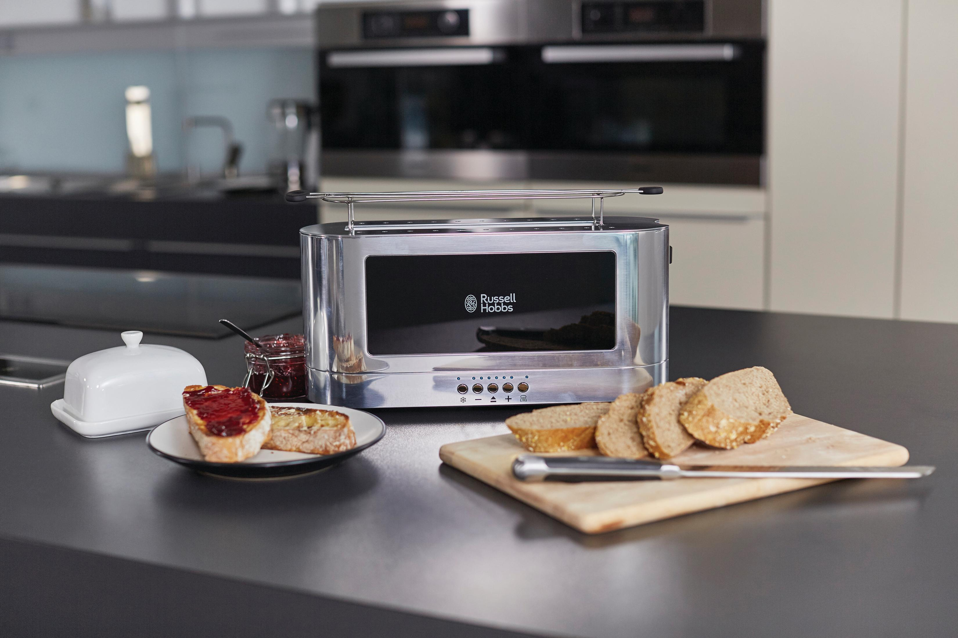 Ein Toaster aus Edelstahl mit Brot und Marmelade auf einer Arbeitsplatte. Auf einem Teller liegen geschnittenes Brot und Marmelade.